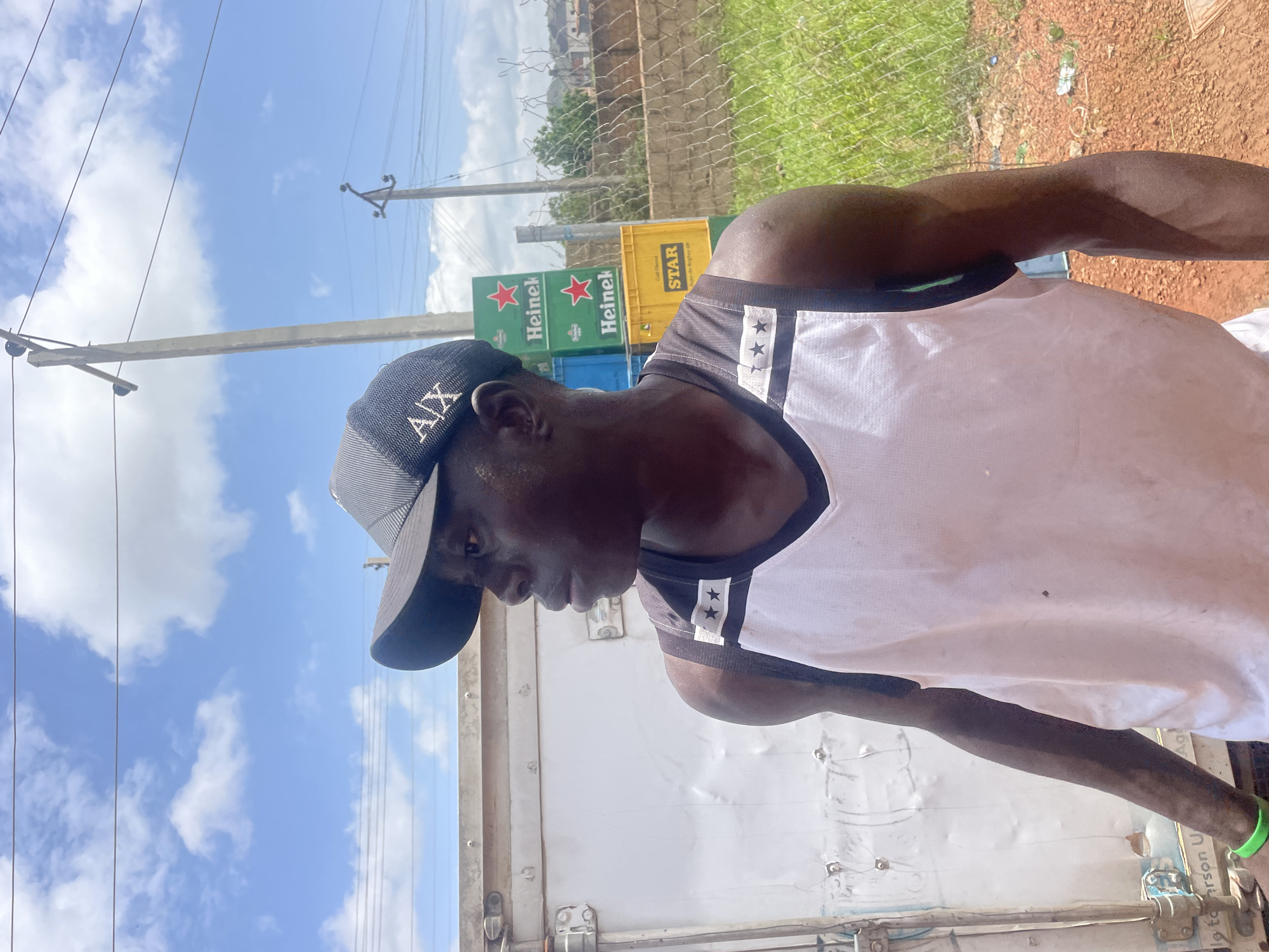 A young man is standing outdoors, facing to his right, with his head turned slightly further. He is wearing a black baseball cap with the letters "AX" in white on the front and a white tank top with black trim and two black stars on the upper chest. He has a green wristband on his left wrist. Behind him, there are stacks of cases of Heineken and Star beer. A white refrigerated truck is to his left, and a chain-link fence with overgrown grass behind it is to his right. Utility poles and power lines are visible in the background under a cloudy blue sky. The ground is covered in reddish-brown soil. The scene appears to be in an open, possibly commercial or storage area, within the city of Gura Top, Nigeria.
