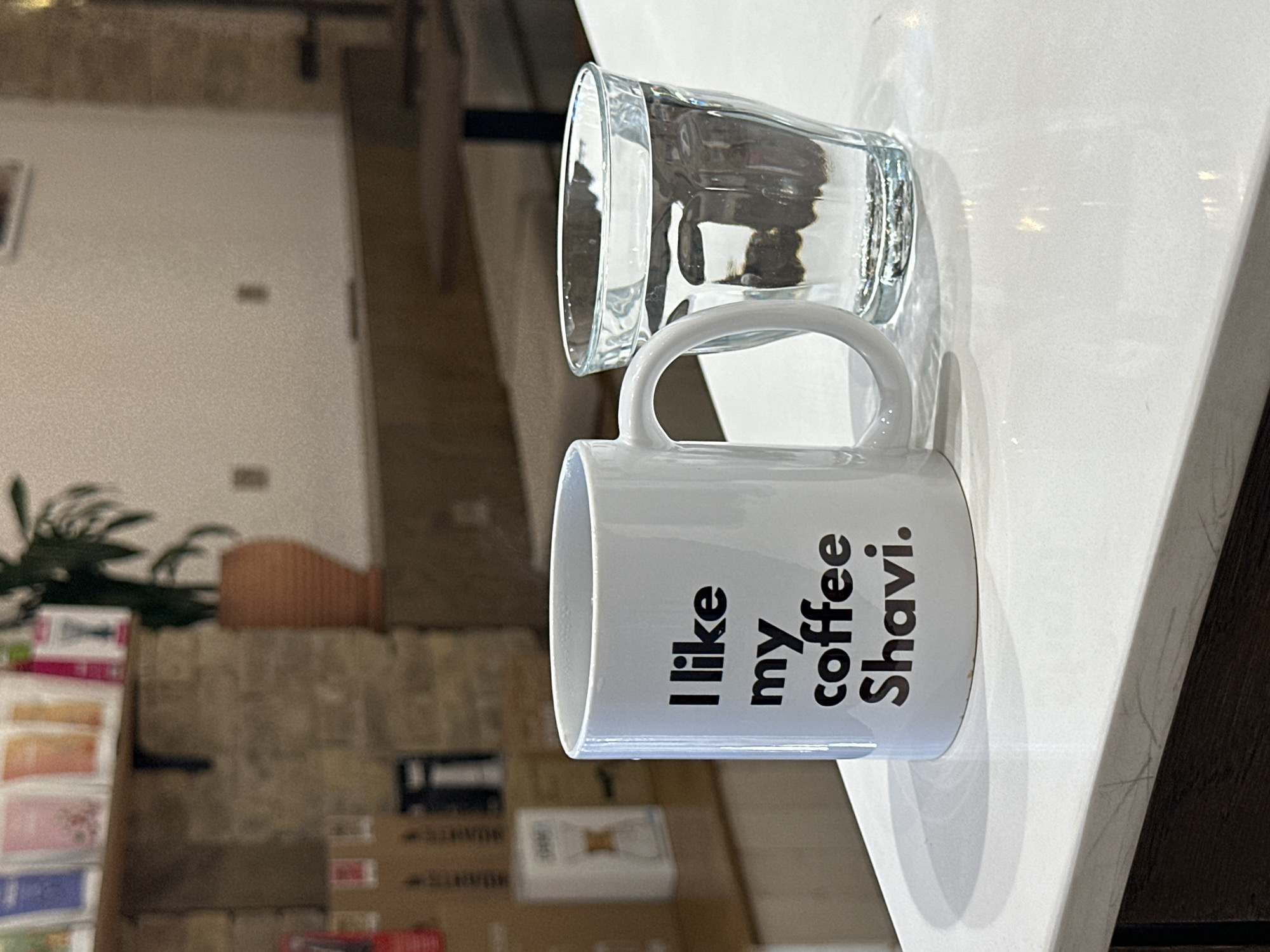 The scene is a close-up shot of a white ceramic mug and a glass of water resting on a white countertop. The mug has black text vertically printed on its side, reading "I like my coffee Shavi." The glass of water sits partially behind and above the mug. In the background, blurred, is a display of packaged goods or merchandise and a plant, suggesting a commercial establishment. The location is Tashkent, Uzbekistan.