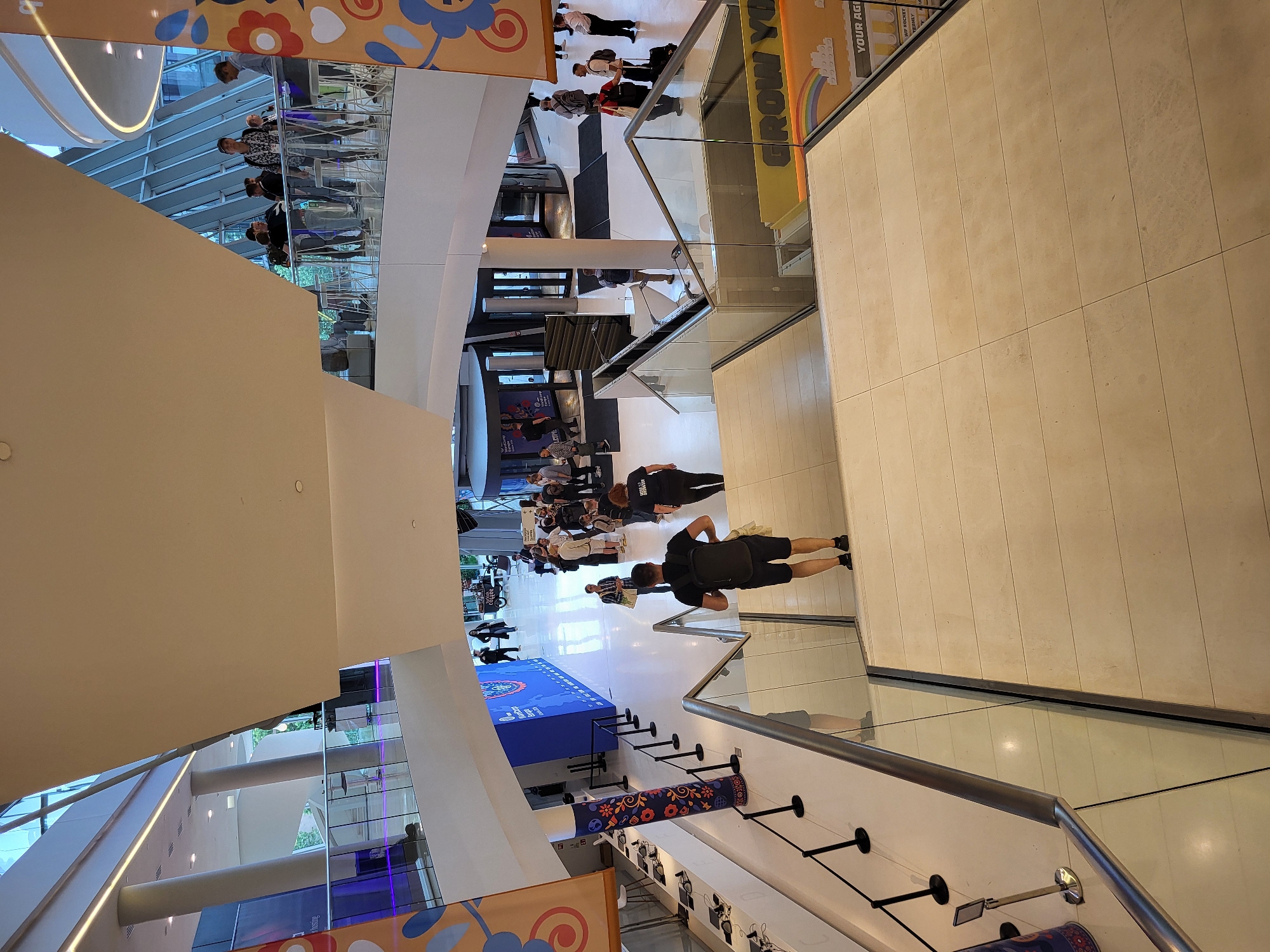 This high-angle shot captures a multi-level, modern indoor event venue. The dominant foreground features a wide, light-tiled staircase with a glass and metal railing, descending from the viewer's perspective. A male figure, seen from behind, wearing a black t-shirt, black shorts, and a backpack, is descending these stairs on the right.
The mid-ground and background reveal a bustling lower level and an upper-level balcony. The lower level features light-colored tiled flooring and a large blue wall on the left with white text that reads "WordCamp Europe WELCOME" and an accompanying logo. Black stanchions form a queue line leading along this wall. Numerous individuals are scattered across the lower level, mostly dressed casually, walking, standing, and conversing. Several groups of people are gathered near large revolving glass doors, likely main entrances or exits, located in the center-right of the lower level. A partially visible sign on the bottom right of the image, below the railing, features a rainbow, block-like graphics, and the text "GROW YOU" and "YOUR AGI".
The upper level, visible on both sides of the image, features a walkway or balcony with similar glass and metal railings. People are visible on this upper level, observing the activity below or interacting with each other. The architecture includes tall, light-colored pillars and large glass window sections that suggest natural light. Large banners hang from the ceiling, one on the left with blue and orange organic shapes, and another on the right with an orange, blue, and red floral/heart pattern. The ceiling itself is high and white, with visible recessed lighting and linear light strips along the upper-level edges. The overall environment is brightly lit, clean, and appears to be a busy conference or exhibition space for a WordCamp Europe event in Unknown, Unknown.