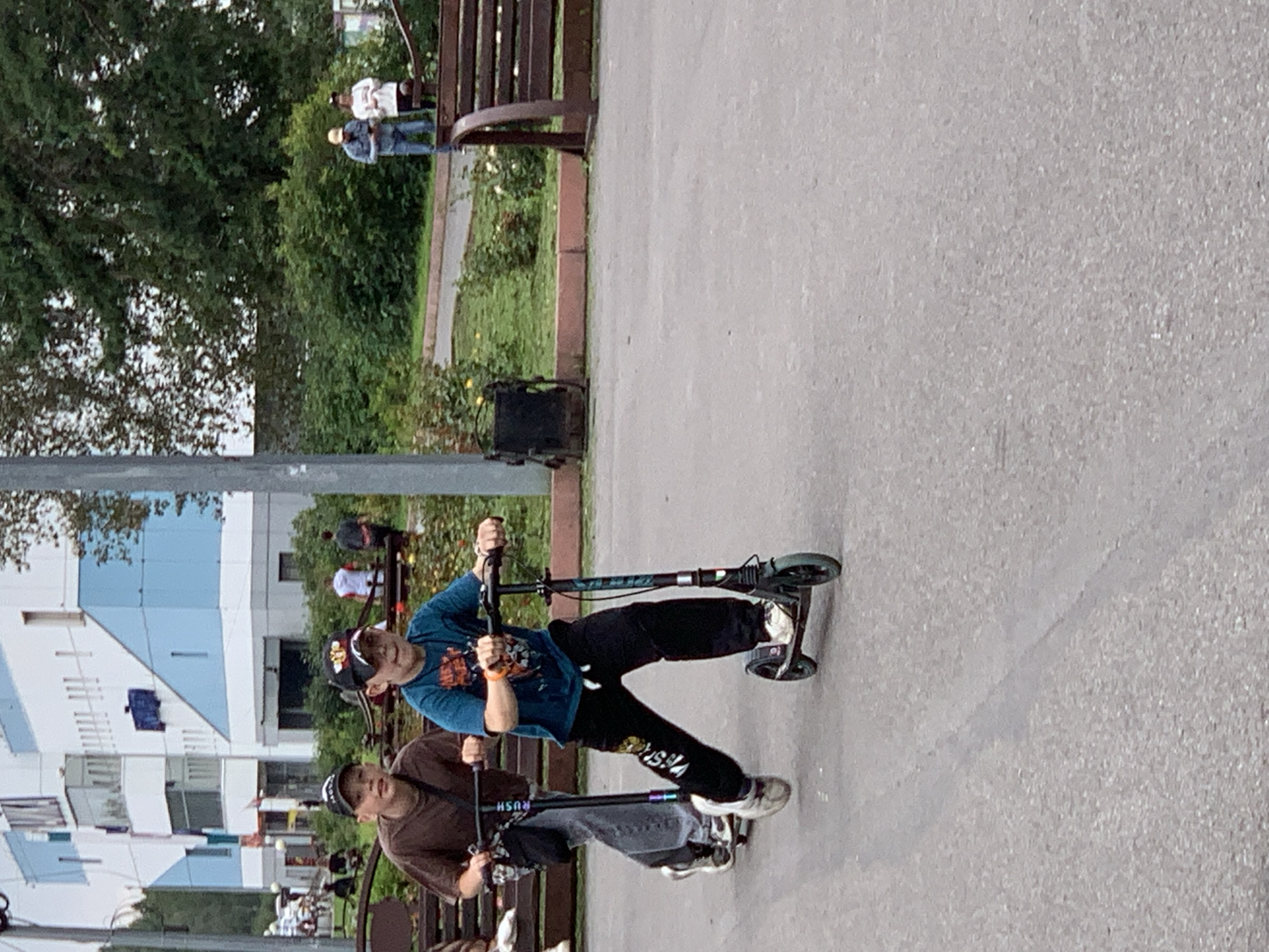 The image depicts two boys riding scooters on an asphalt pathway. The boy in the foreground is wearing a teal t-shirt, black pants with white accents, and a black baseball cap. He is actively riding his scooter, with his body angled forward. The boy behind him is wearing a brown t-shirt, gray pants, and a black baseball cap. He is also on his scooter, appearing to be keeping pace with the boy in front. The setting appears to be a park or recreational area, with benches and trees visible in the background. A modern building with blue and white sections is also present in the distance. The overall atmosphere suggests a casual outdoor activity. It is not possible to definitively determine the location as Prokopyevsk, Russia from the image alone.