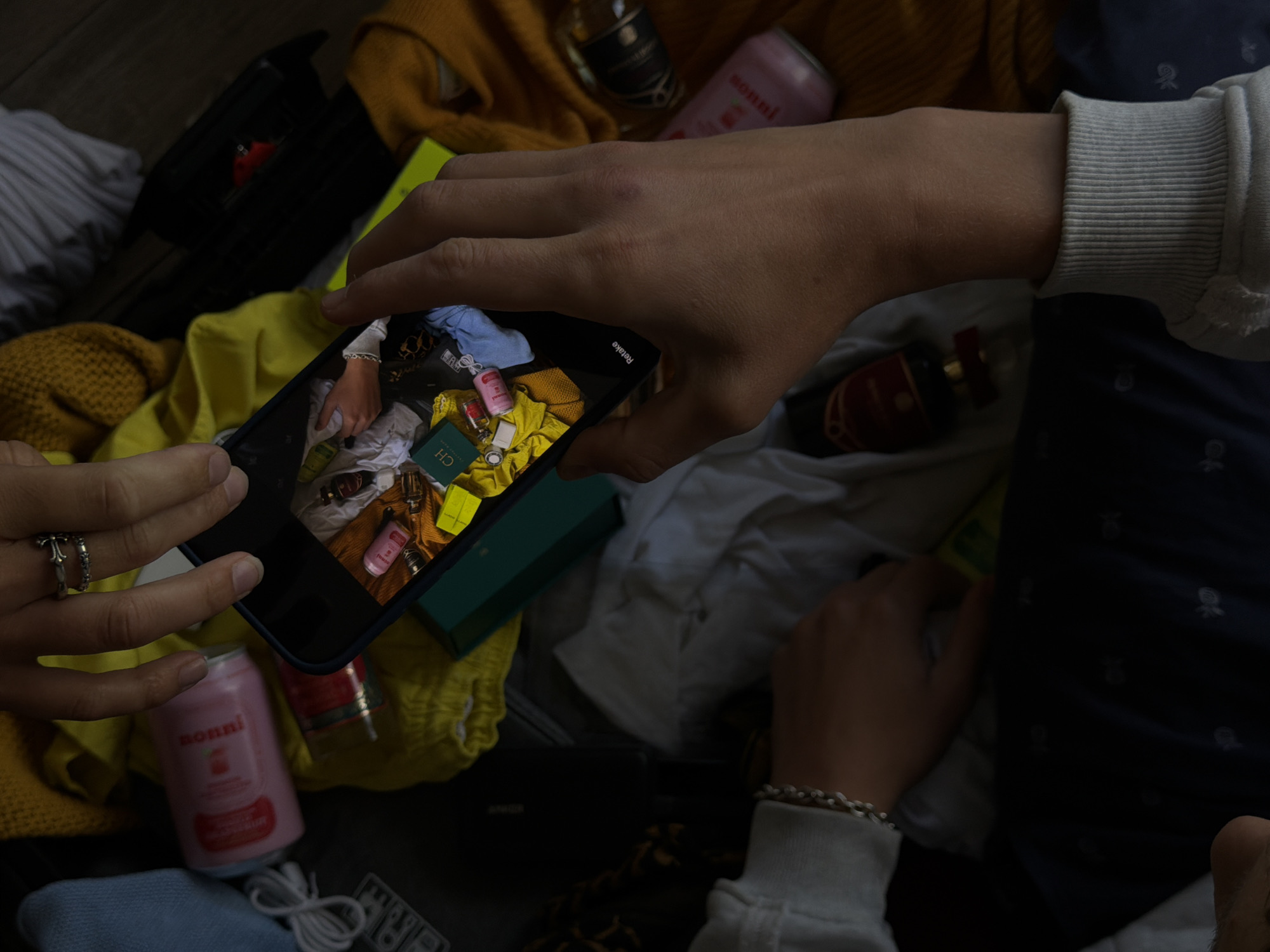 The image shows a top-down view of a person's hands holding a smartphone, which is displaying a photograph of various items. The items in the photograph include a pink can, several small bottles, a green box, and some clothing. In the foreground, another person's hands are visible, one wearing a ring. The background is filled with clothing in various colors, including yellow and blue, and a dark suitcase. The scene appears to be set indoors, on a wooden surface. There is no identifiable information regarding the location in London, United Kingdom.