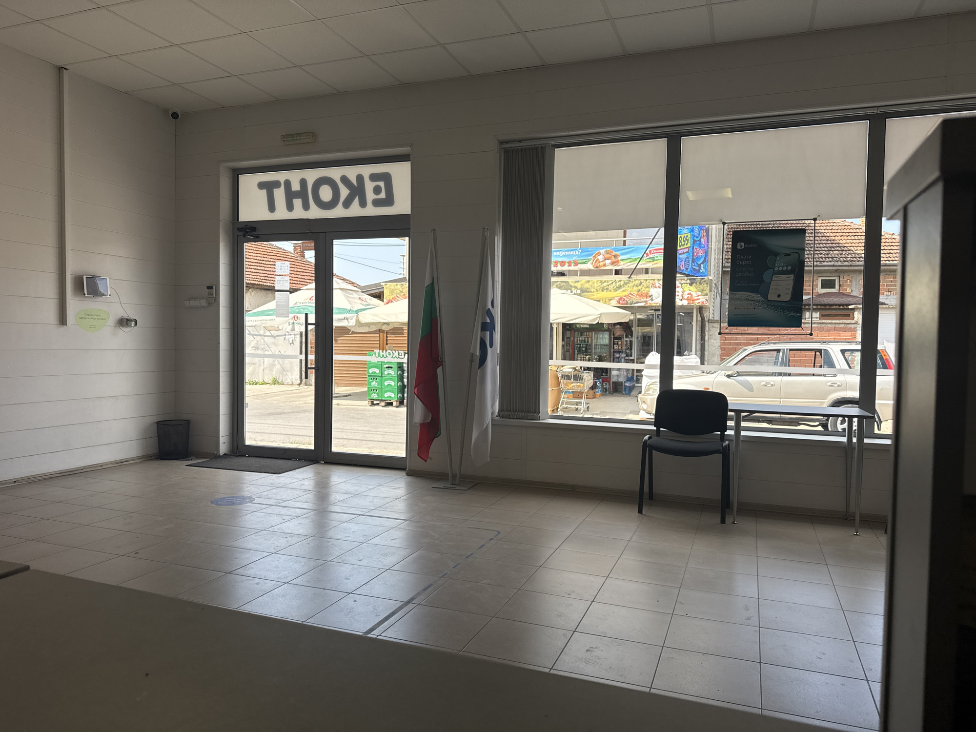 The image shows the interior of a retail establishment, likely a small convenience store or office, located in Rakovski, Bulgaria. A prominent sign with backward lettering, reading "EKON", is visible above a glass entrance door. Inside, the space is tiled and appears sparsely furnished. To the left, a security camera is mounted on the wall, and a small digital display is affixed to the wall below it. Further left, a dark-colored trash receptacle stands near the entrance.
In the center of the entrance area, two flags are displayed on stands: one appears to be the Bulgarian flag, and the other a white flag with blue lettering and a blue logo. Through the glass doors and windows, an outdoor scene is visible, featuring market stalls with awnings, crates of beverages stacked on pallets, and parked vehicles.
A small table with two chairs is positioned by the window on the right side of the interior, looking out onto the street. One chair is occupied by a person. The overall impression is of a functional, if somewhat dated, commercial space.
