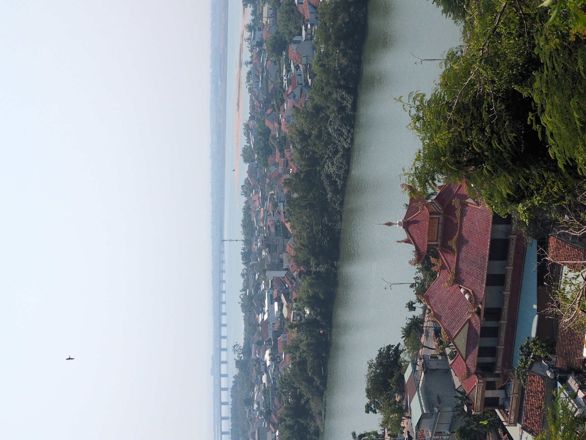 This is an aerial view overlooking the city of Tuy Hòa, Vietnam. The scene contains a river running vertically through the center of the frame. On the river's right bank, a prominent traditional building with a red tiled roof and ornate architecture is visible. The city's skyline with its predominantly red-tiled roofs extends to the left of the river. A long bridge stretches across the horizon. A bird is flying in the pale sky. Trees are visible along the riverbank.