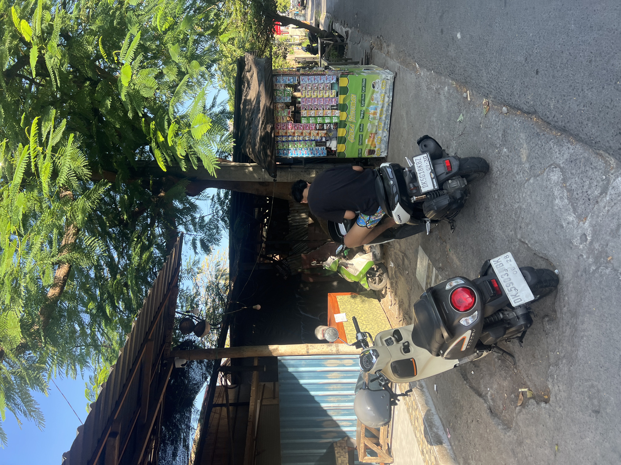 A man sits on a scooter parked on a paved road in Sukawati, Indonesia, next to a small roadside stall selling various beverages. The stall, brightly painted green and yellow, is covered by a black tarp and displays an assortment of packaged drinks and snacks. Behind the stall, a larger, more permanent structure with a corrugated metal roof is visible. The man is wearing a black t-shirt and patterned shorts. Two other scooters are parked nearby, one a light cream color and the other green. The scene is shaded by a large tree with lush green foliage. The road surface is rough and uneven, with some cracks and debris. The sky is clear and blue.