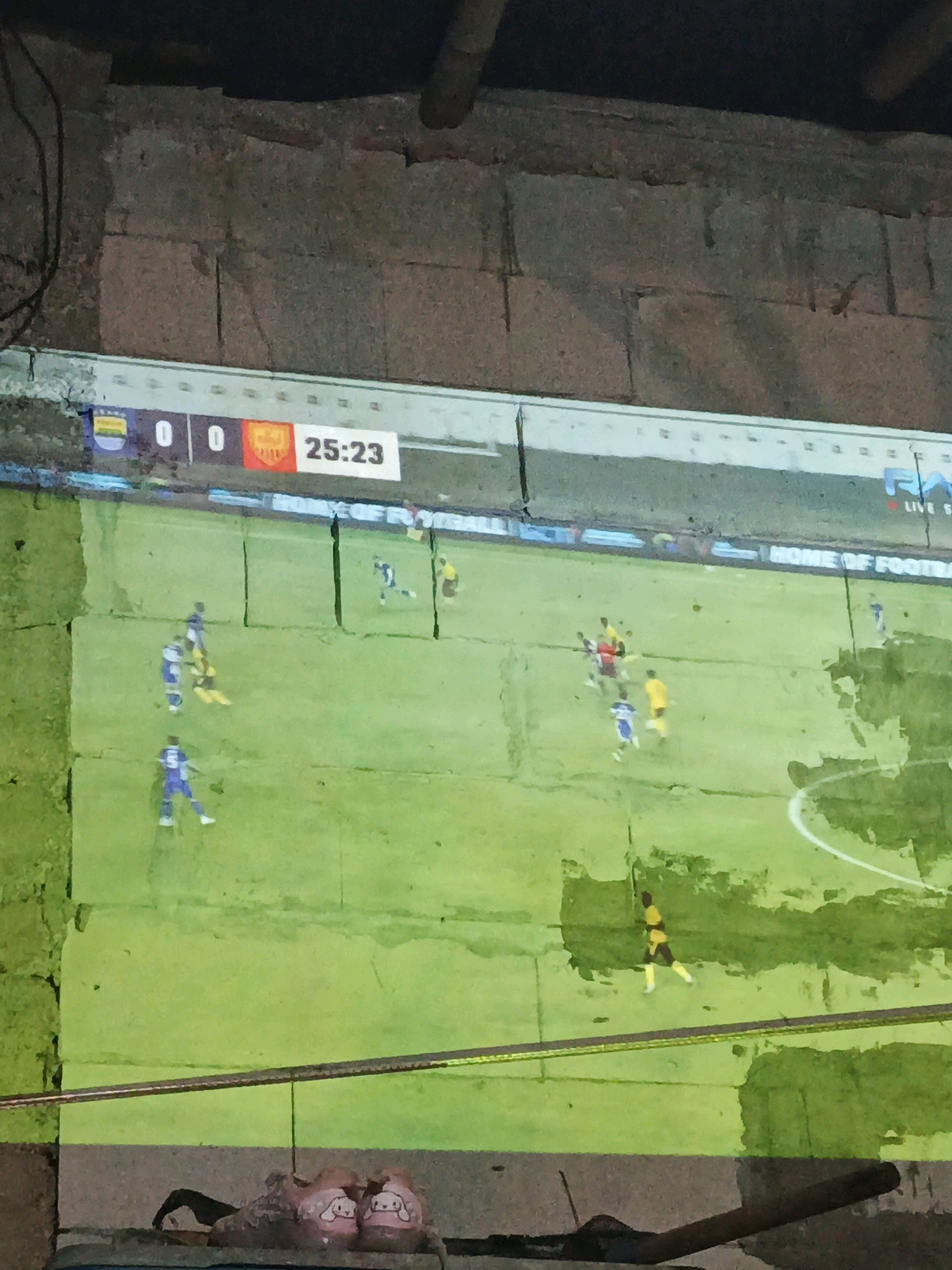 The image displays a projection of a soccer match onto a rough, textured wall. The score displayed on the projection is 0-0 with the game time at 25:23. Several players in blue and yellow uniforms are visible on the projected field. In the foreground, a pair of pink slippers with cartoon characters are placed on a ledge, obscuring a portion of the projection. The scene appears to be outdoors, possibly in a courtyard or alleyway, given the visible brickwork and overhead pipe. The location is identified as Tasikmalaya, Indonesia.