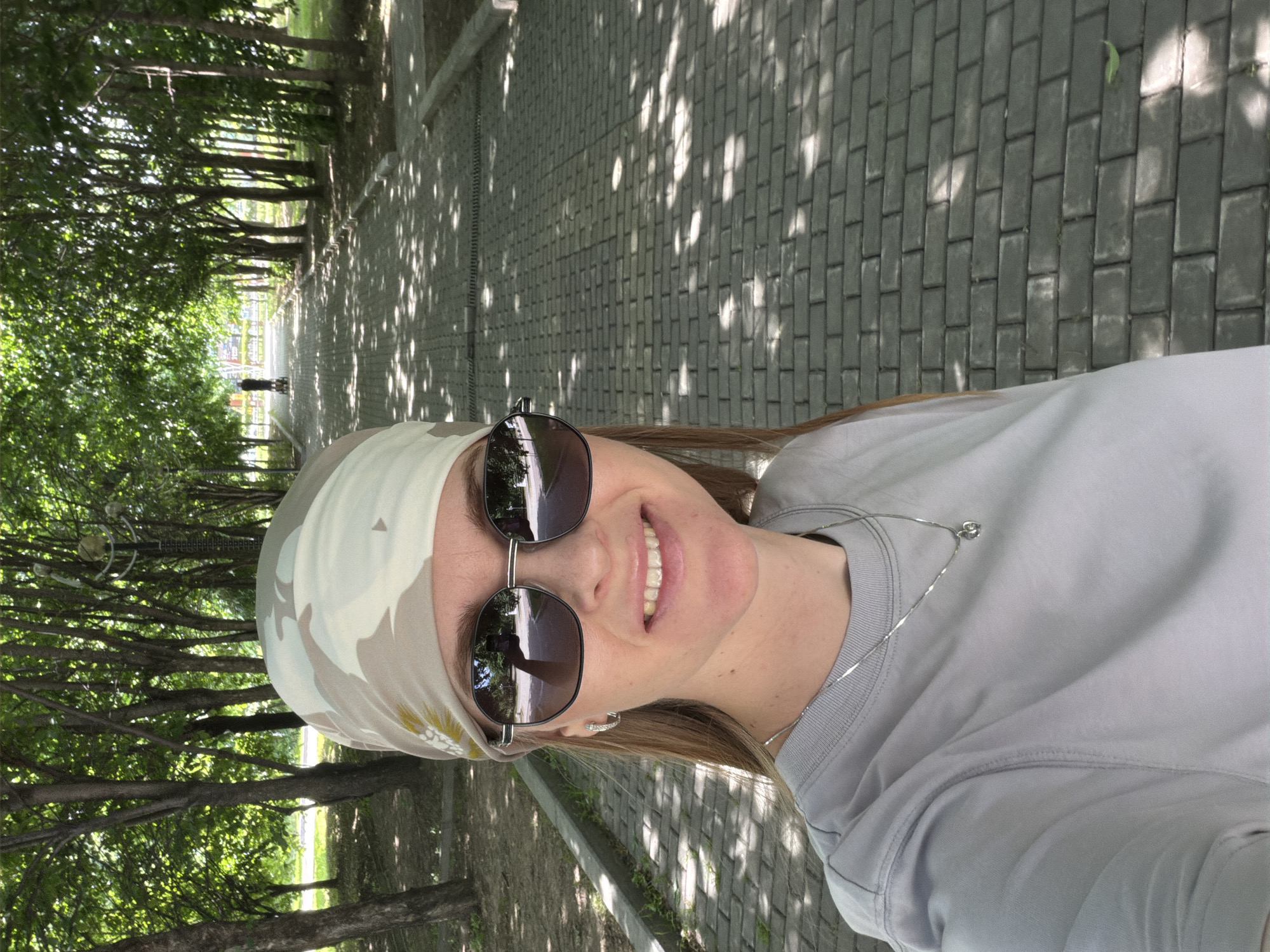 In Khabarovsk, Russia, a woman is taking a selfie on a paved park path lined with trees. She is wearing a camouflage bandana, sunglasses, a grey t-shirt, and a silver necklace with a heart pendant. The path is bathed in dappled sunlight, creating a pattern of light and shadow. In the background, another person is visible in the distance, pushing a cart. The trees suggest a natural or park setting within the city.