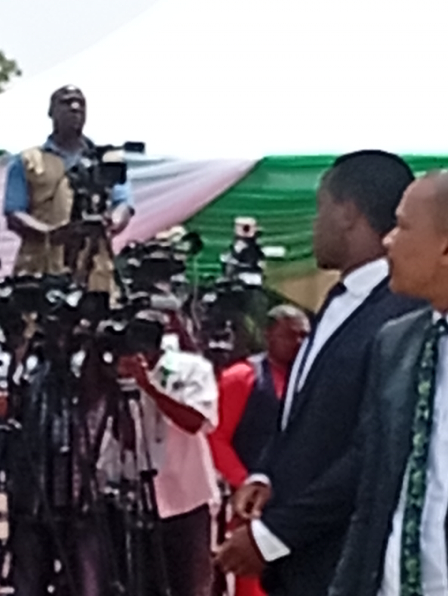 This image captures an outdoor media event or public gathering in Unguwan Sanusi, Nigeria, likely during the daytime under bright, overcast skies.
In the foreground, a group of men is visible. On the right, two men are seen from the chest up, facing right. The man closer to the viewer has light brown skin and is partially visible, wearing a suit jacket and a tie with a patterned design, over a white shirt. Next to him, another man with dark skin is seen in profile, wearing a dark suit with a white shirt and a dark tie or bow tie.
To the left and in the mid-ground, numerous professional video cameras on tripods are set up, indicating a significant media presence. Several cameramen are operating these cameras. One cameraman in the upper left section is standing, wearing a light blue shirt and a tan or light brown vest, holding a camera. Further back, another cameraman is crouched or bending over a camera.
The background features elements of an event setup, including what appears to be a light-colored tent or canopy with green and pink/red fabric backdrops or decorations. The overall scene suggests a formal occasion, possibly a press conference, rally, or official ceremony, given the media coverage and the attire of some attendees. No specific text is clearly visible in the image.