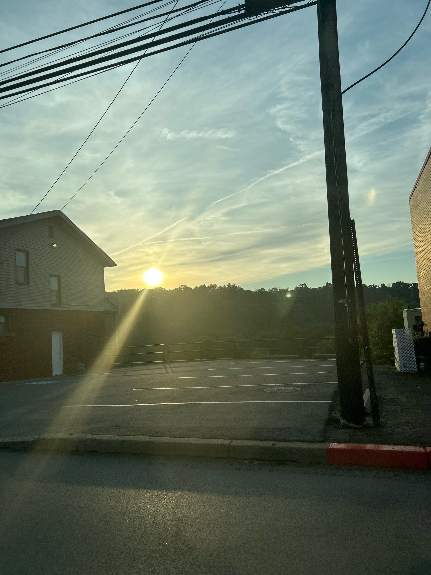 This photograph, captured in Collier Township, USA, depicts a street-level view at sunrise or sunset. The sun, a bright orb low in the sky, casts long rays of light across the scene. In the foreground, a paved road with a curb painted red at the far right is visible, with a section of asphalt parking lot marked with white lines further back. To the left, a two-story building with beige siding and dark brick lower level is situated. A white door is present on the brick portion. A tall, dark utility pole stands prominently in the right half of the frame, with numerous overhead wires stretching across the upper portion of the image against a light blue sky streaked with wispy clouds and contrails. Beyond the parking lot, a densely wooded hillside forms the backdrop, silhouetted against the bright sky. The overall impression is one of an urban or suburban edge transitioning into a natural landscape during the golden hour.