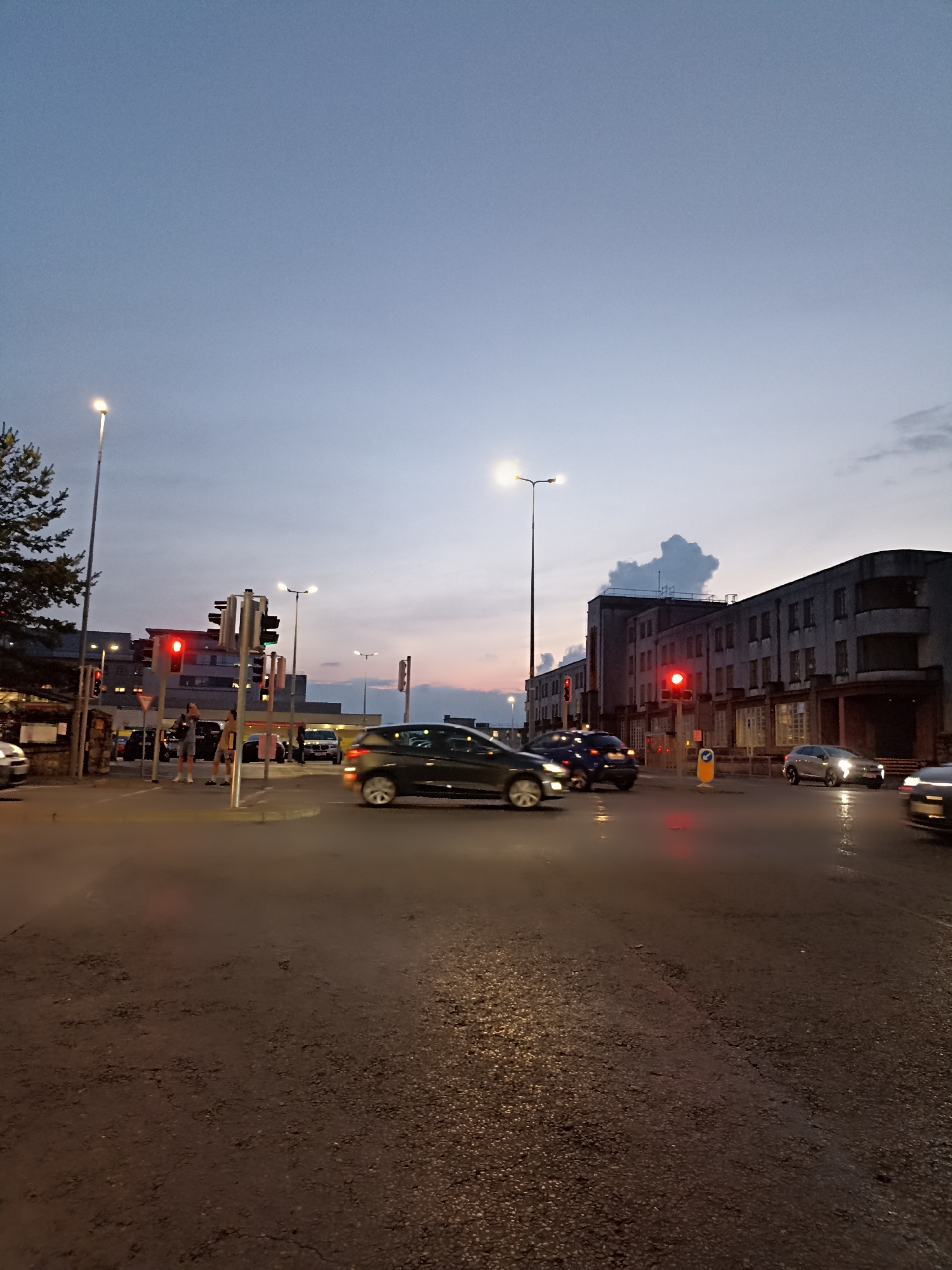 This image depicts a street scene at dusk, likely in the city of Galway, Ireland, given the architectural style and the context of the provided image. A busy intersection is captured, with several cars navigating the wet road. Traffic lights are visible, with one clearly showing a red signal. Two individuals stand near a traffic light pole on the left side of the frame, one holding a phone to their ear. In the background, a multi-story building with a distinctive facade lines the street. The sky is a blend of twilight hues, with hints of pink and orange near the horizon, suggesting the sun has recently set. Streetlights cast a warm glow on the wet pavement, reflecting the lights of the vehicles and creating a somewhat atmospheric, urban environment. The overall impression is that of a typical evening in a European city, with everyday activities like driving and walking taking place.