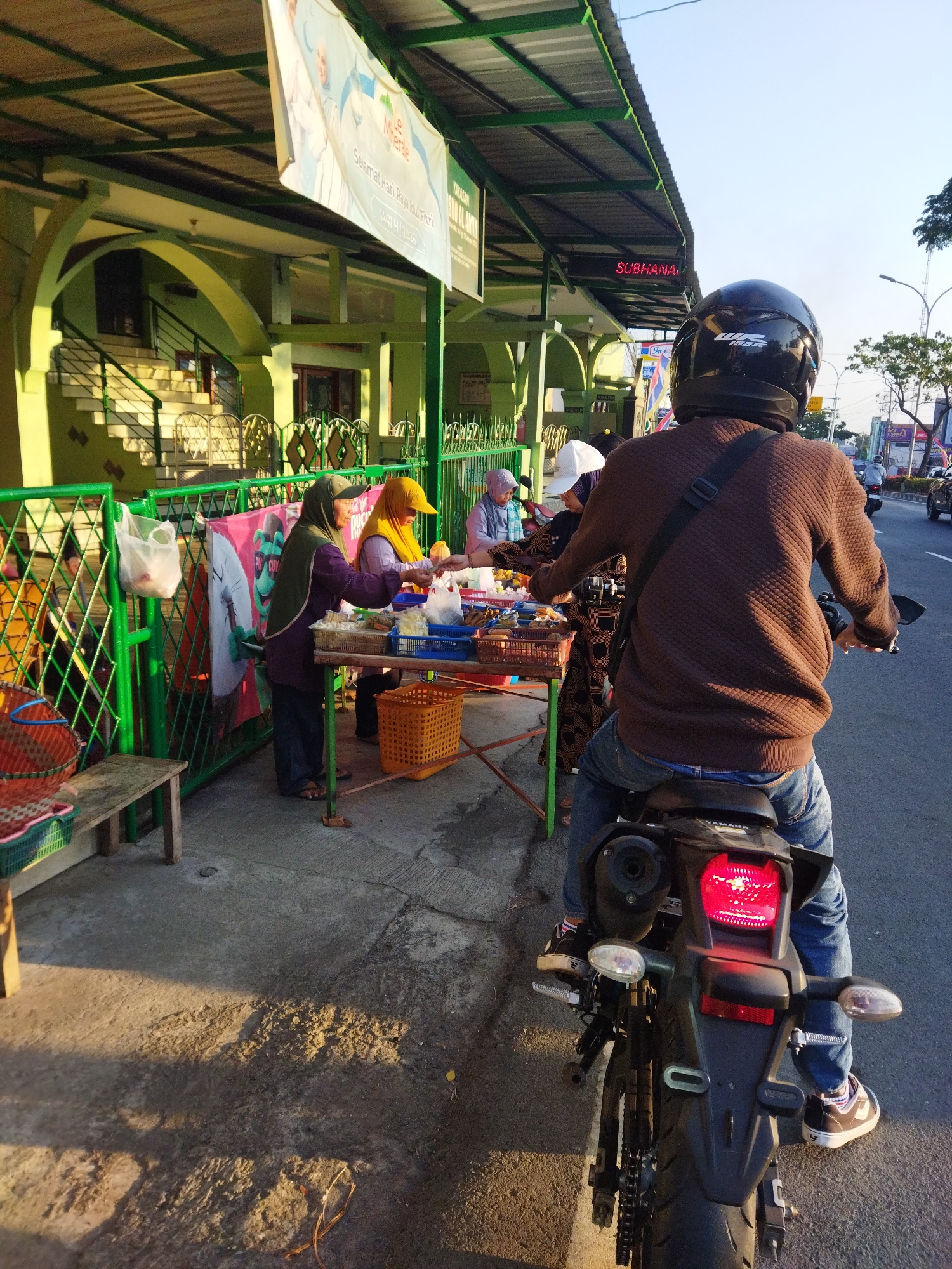 A motorcyclist, viewed from behind, is stopped next to a street-side market stall. The motorcyclist wears a brown quilted sweater, blue jeans, and a black helmet with a white logo. The motorcycle's brake light is illuminated. The market stall is set up under a green awning extending from a building with green arched columns and a yellow staircase. Several women are attending the stall, arranging food items. One woman wears a green hijab and a purple top, another a yellow hijab and a light blue top, and a third a white head covering and a patterned dress. The stall displays various packaged food items and baskets. A green metal fence separates the market from the sidewalk. A banner displaying text in Indonesian hangs above the market. In the background, a street is visible with other motorcycles and cars. The scene is illuminated by sunlight, casting long shadows on the ground, indicating it is likely late afternoon or early morning. The building and street signage suggest an urban setting, possibly in Semarang, Indonesia, given the presence of local market practices. A digital sign above the market reads "SUBHANA" in red letters.