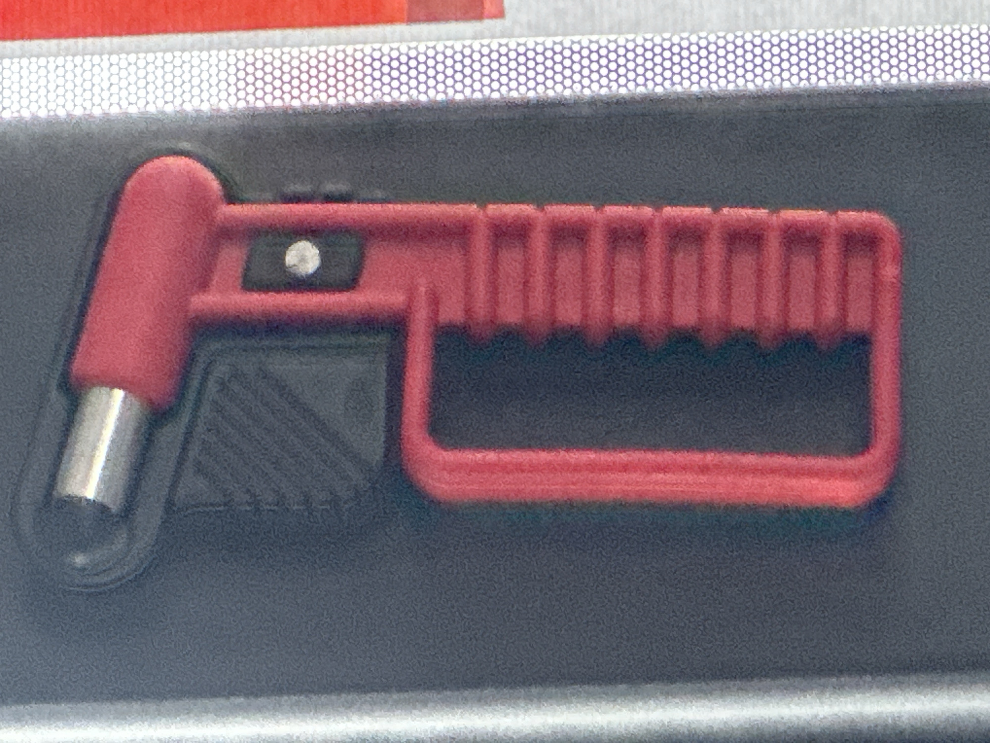 A red and silver car safety hammer is attached to a dark gray surface. The hammer has a pointed silver metal end designed for breaking glass, and a red plastic handle with a grip pattern. A small silver button is visible on the side of the handle. The dark gray surface appears to be a textured material. To the right of the hammer, there is a strip of reflective material with a pattern of small white circles on a gray background, and a further red strip. The overall image is a close-up, likely taken within a vehicle, possibly in the city of Günzburg, Germany.