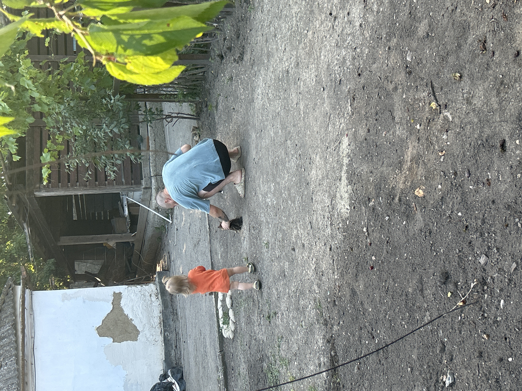 In Prokhladny, Russia, a man in a light blue t-shirt and shorts is crouching on a dirt path, interacting with a small black and white kitten. Beside him, a young child wearing an orange dress stands, observing the scene. The ground is a mix of dirt and small stones. Behind the man, a rustic wooden structure, possibly a shed or an outbuilding, is partially visible, with some trees and greenery nearby. To the left, a white-painted wall with patches of exposed plaster suggests a weathered building. The lighting indicates it is daytime, likely late afternoon or early evening, with the sun casting shadows. The overall scene depicts a casual outdoor moment between people and an animal in a rural or semi-rural setting.