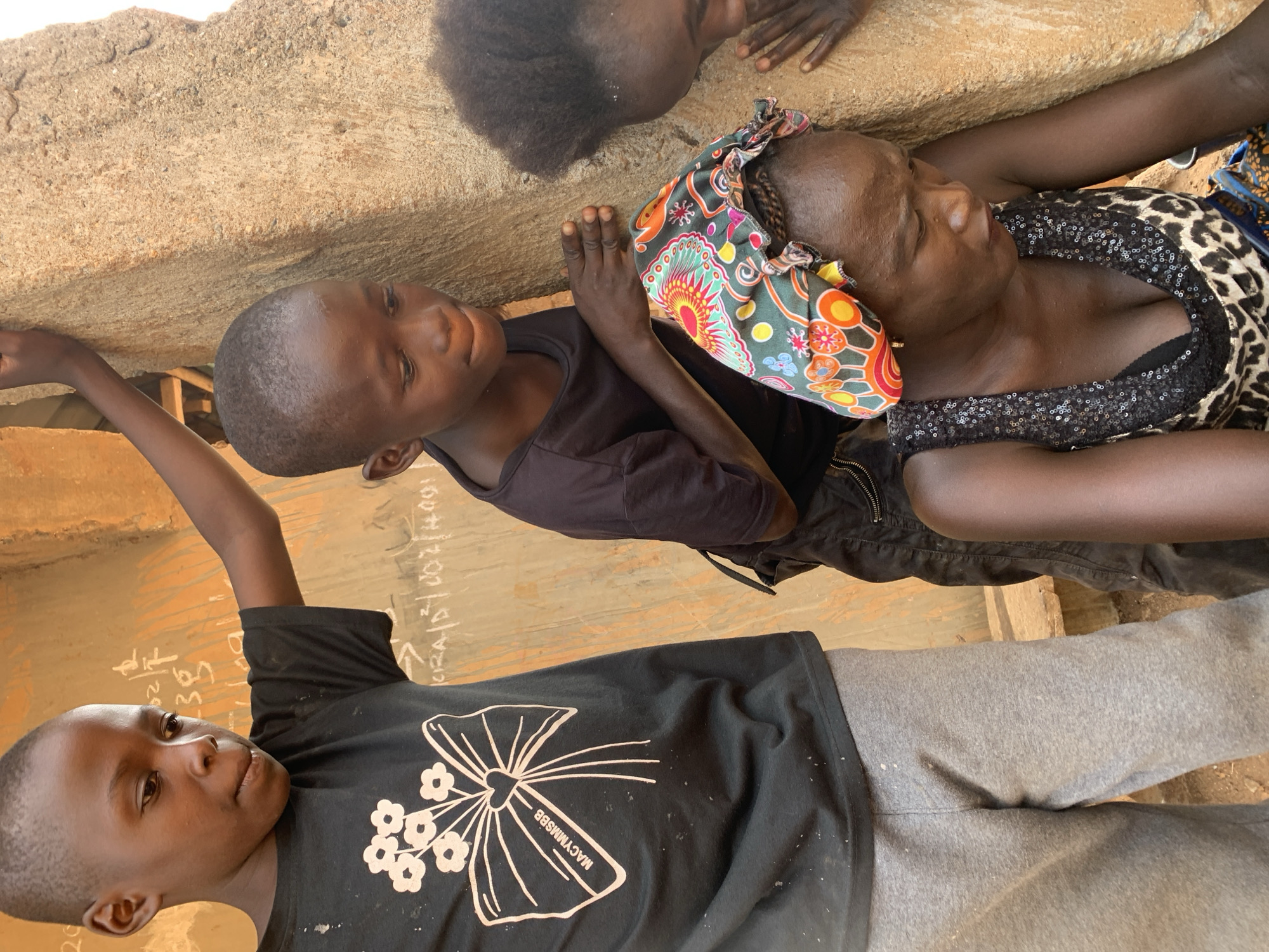 The image depicts three individuals, possibly in Kukum, Nigeria, given the prompt's instruction. One is a woman with dark skin, wearing a leopard-print top with sequined detailing and a colorful headscarf. She is positioned near the top of the frame, lying down. A young boy with dark skin, wearing a black t-shirt, is next to her. Another boy, also with dark skin, wearing a black t-shirt with a white floral design, and grey pants, is lying down near the bottom of the frame. The backdrop appears to be a wall or surface with some writing on it. The scene appears informal and relaxed.