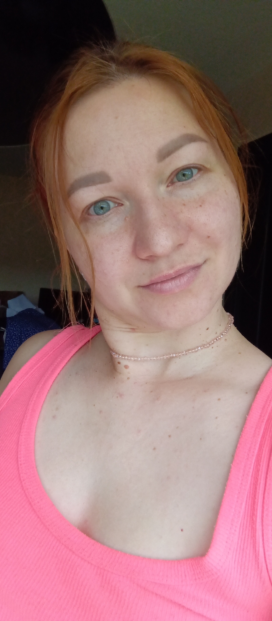 A close-up portrait shows a fair-skinned woman with freckles and red hair, wearing a pink tank top and a delicate pink beaded necklace. Her eyes are a striking blue-green color. The background is partially obscured, revealing a glimpse of a dark surface and a piece of blue and white fabric. The image location and city cannot be determined.