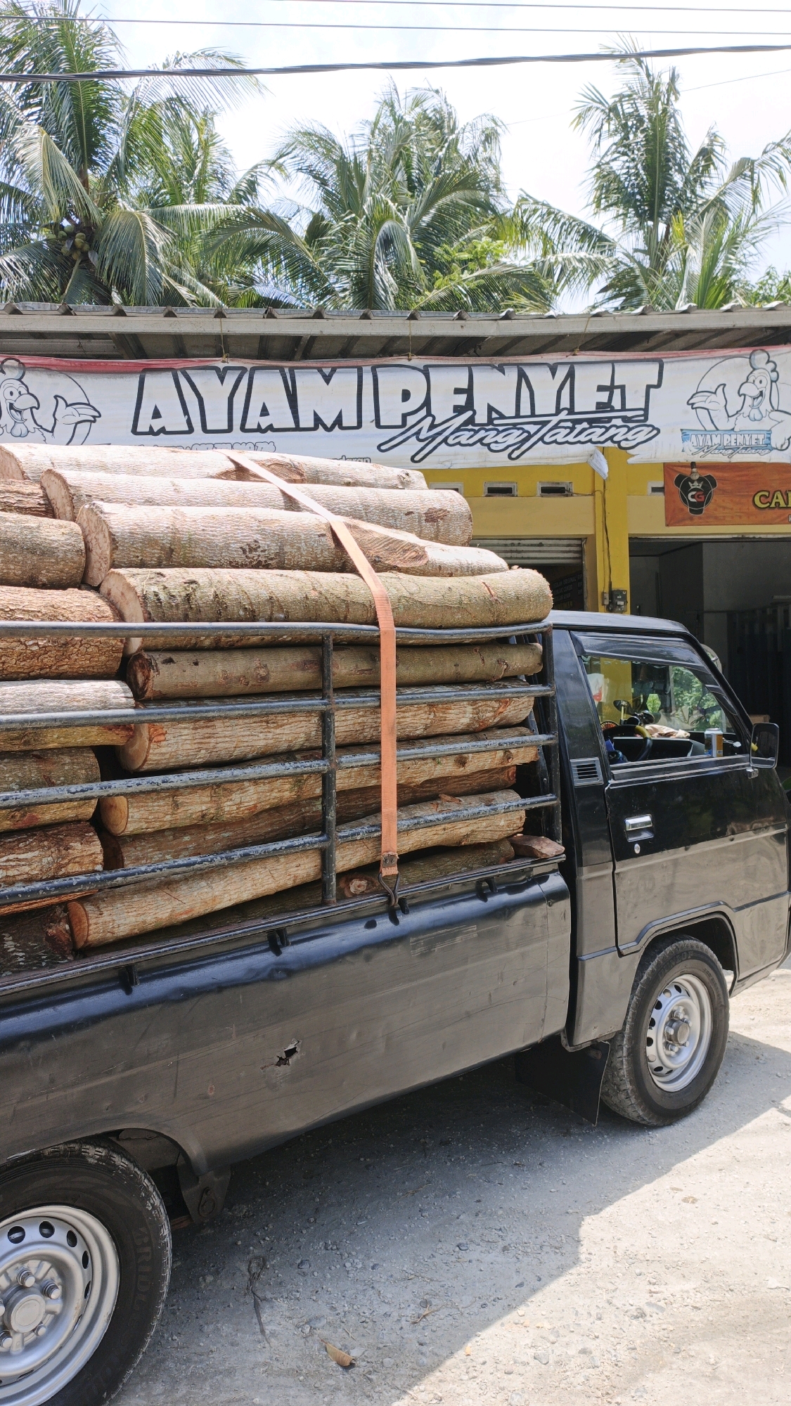 A black pickup truck is parked on a gravel surface in front of a yellow building with a banner that reads "AYAM PENYET". The truck is loaded with a large stack of cut logs, secured with an orange strap. Palm trees are visible in the background, suggesting a tropical or subtropical location. The scene appears to be outdoors, possibly on a sunny day, given the shadows. The building facade shows some windows and signage, including one with a cartoon chicken. The overall environment suggests a commercial or roadside setting in a place like Cibenda, Indonesia.