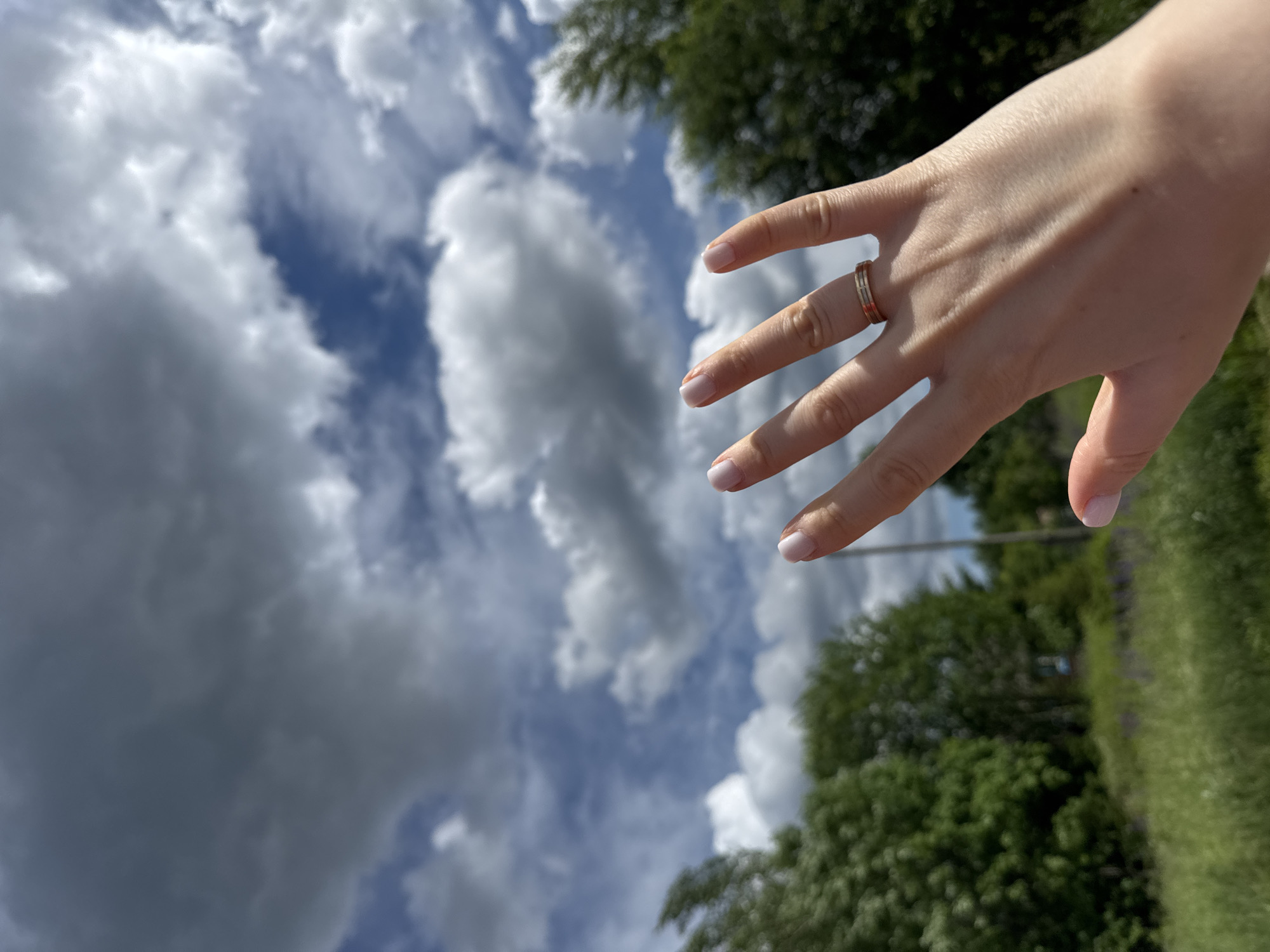 A close-up, slightly angled shot shows a person's left hand, palm facing forward and fingers splayed upwards, in the foreground. The hand has light skin and neatly manicured fingernails painted a pale pink. A thin ring with a striped pattern in gold and possibly another metal is worn on the ring finger. The background is out of focus, featuring a sky with dramatic, dark grey storm clouds interspersed with patches of blue sky and fluffy white clouds. Below the sky, a line of lush green trees and vegetation is visible, with hints of purple flowers in the lower portion of the frame. The overall impression is of a hand reaching towards a dynamic sky, set against a natural, outdoor environment. The image does not provide any specific clues to its location in the city of Chelyabinsk, Russia.