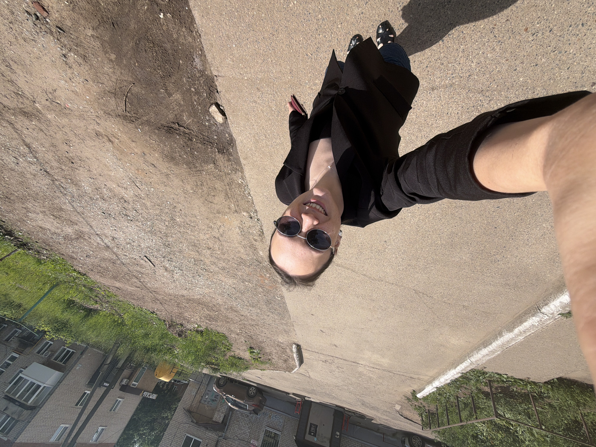 A high-angle, full-body selfie of a woman wearing sunglasses and a black blazer over a black top and jeans, standing on a concrete path in Chaikovsky, Russia. She is holding a red wallet or phone in her right hand. The background shows a dirt path and a brick building with balconies and windows. A car is parked near the building, and grass and a metal railing are visible. The overall scene depicts an urban outdoor environment on a sunny day.