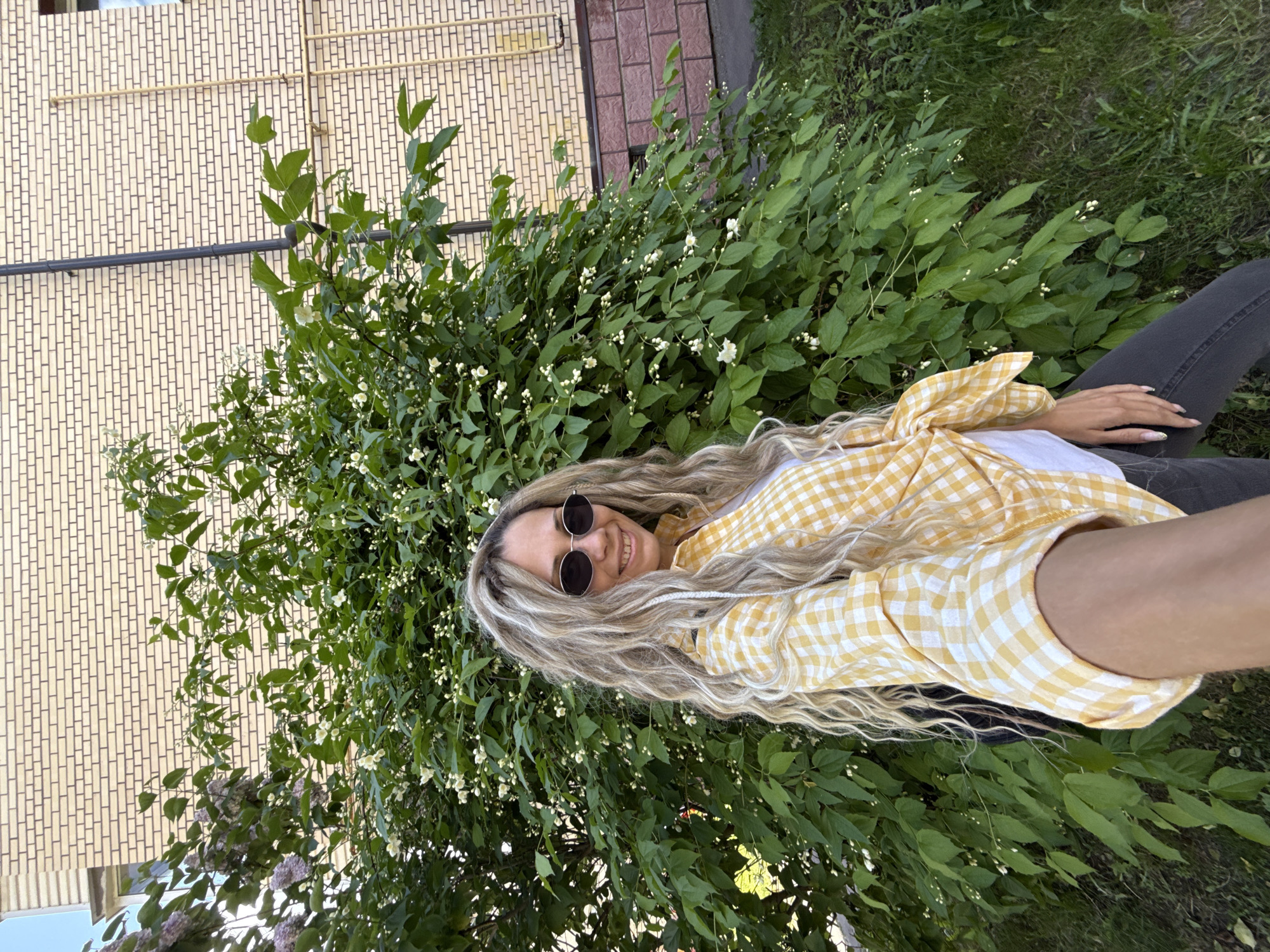 An outdoor, slightly tilted photograph captures a smiling woman against a backdrop of verdant foliage and a building in Rusanovo, Russia. The woman, with long, wavy blonde hair, is wearing dark sunglasses and a yellow and white checkered button-up shirt over a white top, paired with dark pants. Her hand, featuring painted nails, is visible on her leg.
She is positioned in front of a dense green bush laden with numerous small white blossoms, likely jasmine, and a smaller cluster of purple lilac flowers is visible in the lower left. Behind the greenery, a light brown brick building with a distinct pattern of vertical and horizontal lines forms the upper background. Several pipes, one black and others yellow, run along the building's facade. The ground appears to be covered in grass. The scene suggests a bright, sunny daytime setting, and the woman's expression conveys happiness. There is no visible text in the image.