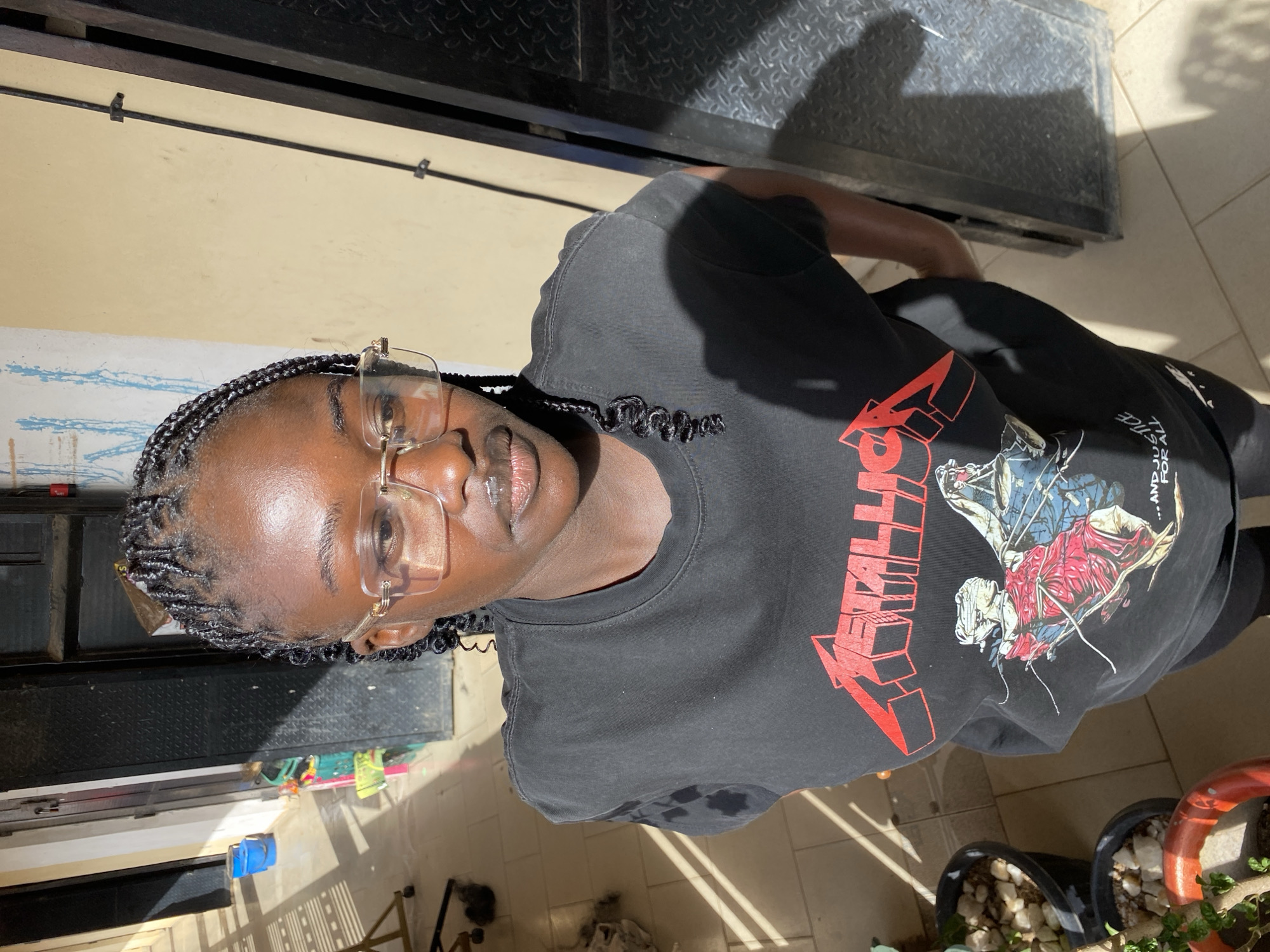 A person wearing glasses and a black Metallica T-shirt with red lettering is photographed from an overhead angle. The subject's hair is braided, with decorative curled edges near the face. The photo is taken outdoors in bright sunlight, casting shadows on the subject and the ground. Several potted plants are visible in the lower right corner of the frame. The location is implied to be in Bukuru, Nigeria.