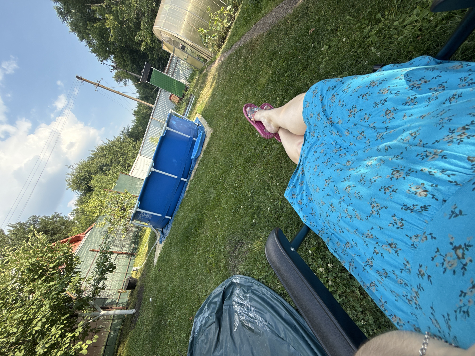 A person, partially visible from the waist down, is sitting in a dark chair in a backyard. They are wearing a blue floral dress and pink flip-flops. The person's legs are crossed, with their feet resting on the grass.
The scene is a sun-drenched backyard with lush green grass. To the left of the person, a blue inflatable swimming pool is set up. In the background, there are trees, a greenhouse, and what appears to be a fence or a building with a corrugated metal roof. A utility pole with wires is visible against a clear blue sky with scattered white clouds.
The time of day appears to be daytime, likely afternoon, given the bright sunlight casting shadows. The weather seems warm and pleasant. The location is suggested to be Nizhne-Chufichevo, Russia, indicated by the presence of a greenhouse and typical backyard elements. The overall mood seems relaxed and peaceful, suggesting a leisurely day spent outdoors. No visible text is present in the image.