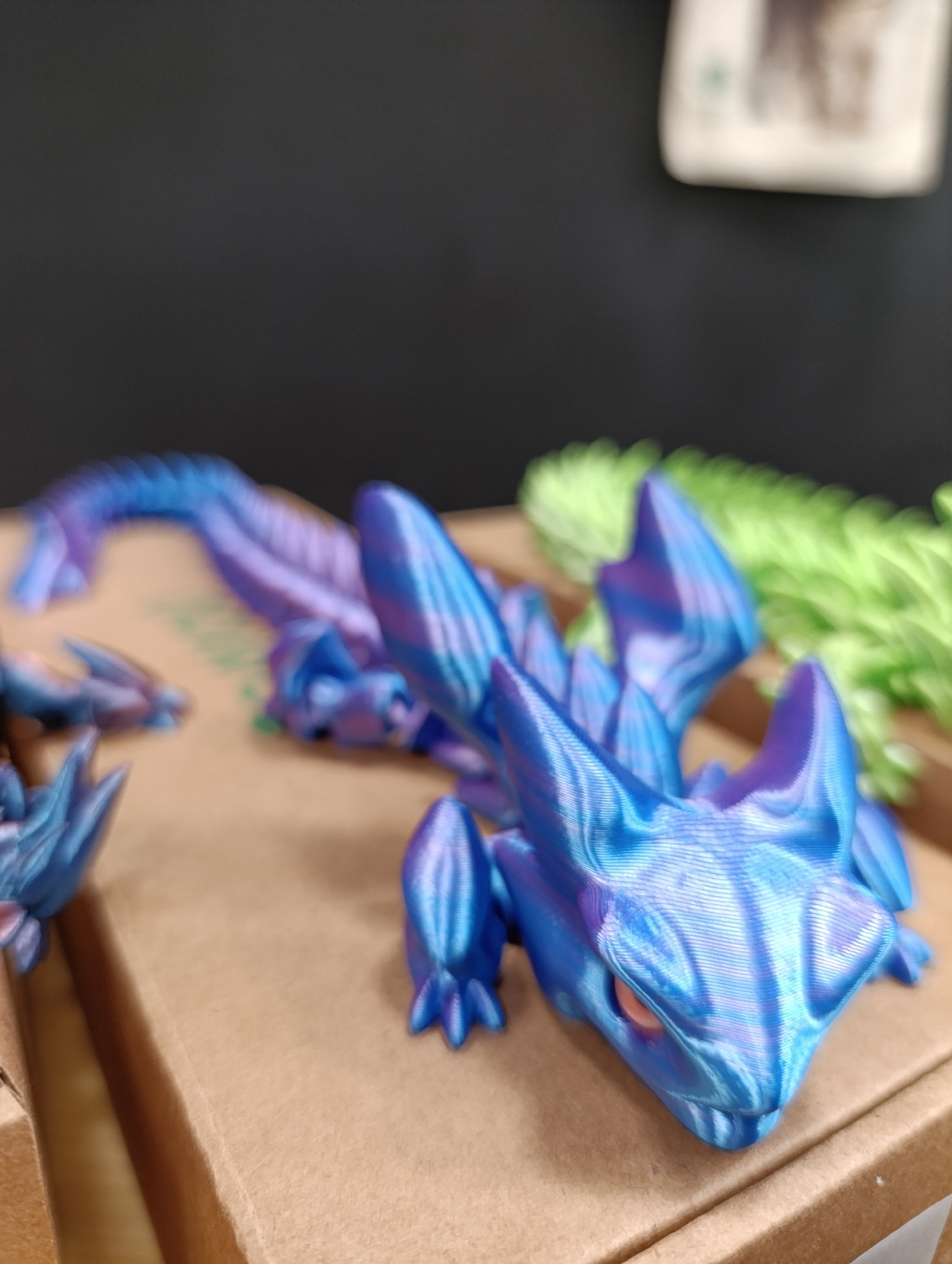 The image shows a collection of 3D-printed articulated dragons on a cardboard surface. The dragons are predominantly blue and purple with a distinct striped pattern caused by the additive layering of the 3D printing process. The main dragon in focus has an orange eye and small feet. The environment is a room with a dark gray background. A blurred green object is visible near the dragon's body. Another partially visible dragon is located to the left. There is a blurry object in the background that is out of focus. The location is unknown.