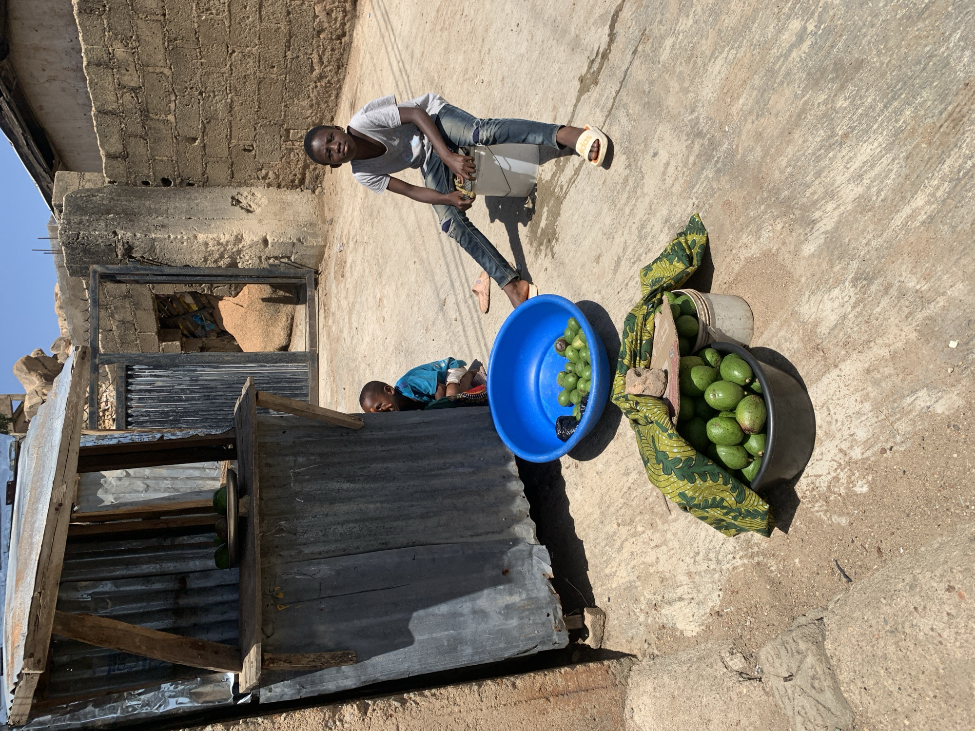In the city of Jos, Nigeria, a daylight scene shows two people and an array of objects related to a possible fruit or vegetable market. A young person is sitting on a white plastic bucket, looking directly at the camera. The individual has short hair, and is wearing a light-colored shirt and blue jeans. A second young person is lying prone on a corrugated metal structure or shelf, facing the same direction as the first individual. A large blue bowl is filled with green produce, situated between the corrugated structure and a bucket filled with more of the same green produce. A patterned green and gold cloth is partially draped over the second bucket. The ground is a light-colored, likely concrete or stone, surface. In the background there is a brick building structure.