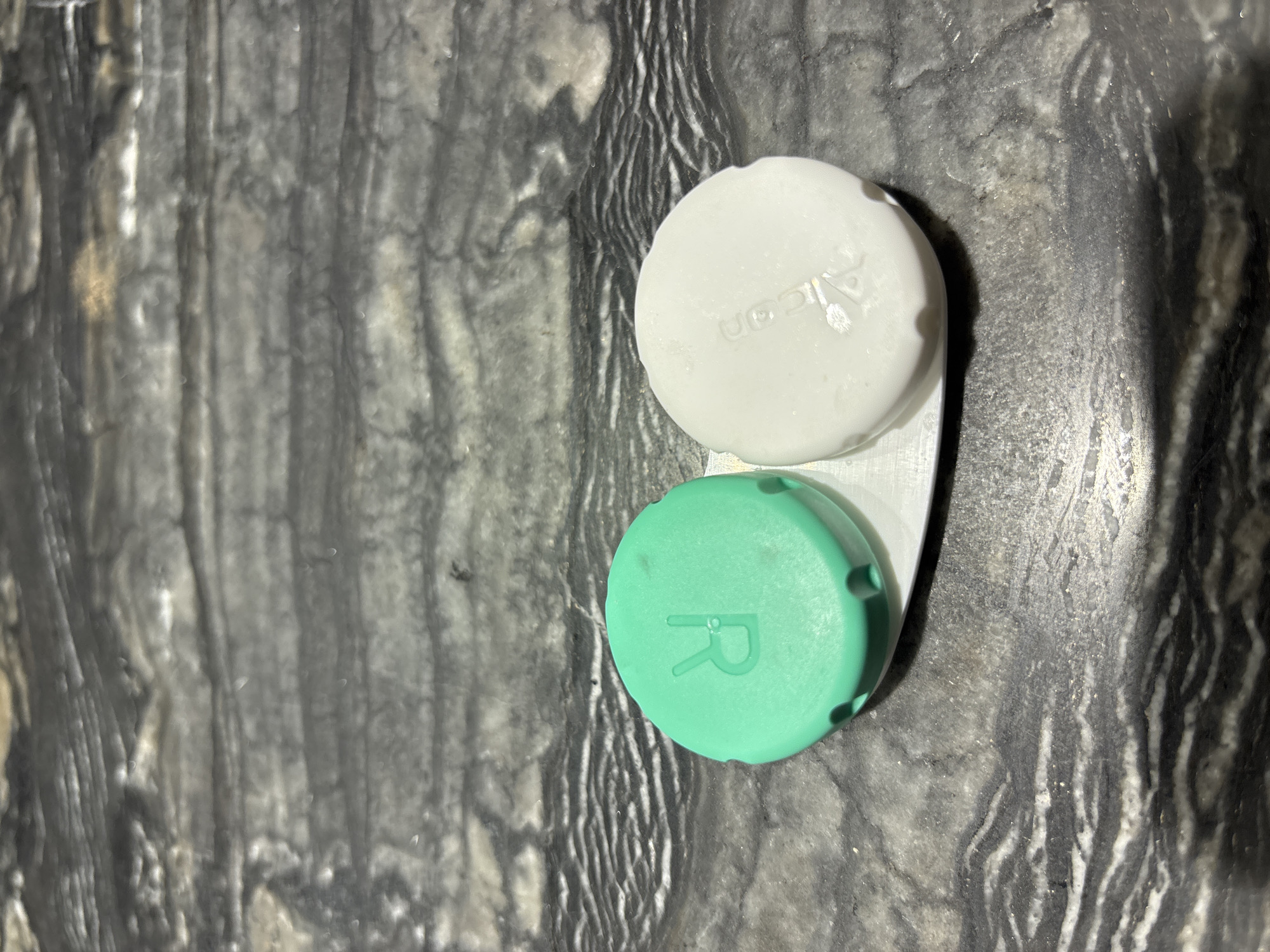 A close-up, overhead shot shows a two-compartment contact lens case against a dark, marbled surface. The case is silver and appears to be metallic. One compartment is topped with a green lid that has a raised "R" marking on it. The other compartment has a white lid with a faint, embossed logo that is difficult to discern but appears to be "Alcon". The marbled surface has prominent dark gray and white striations, suggesting a stone countertop. The scene is lit from above, casting subtle shadows. There are no people, animals, or any indication of the city of Dubai or the United Arab Emirates present in the image.