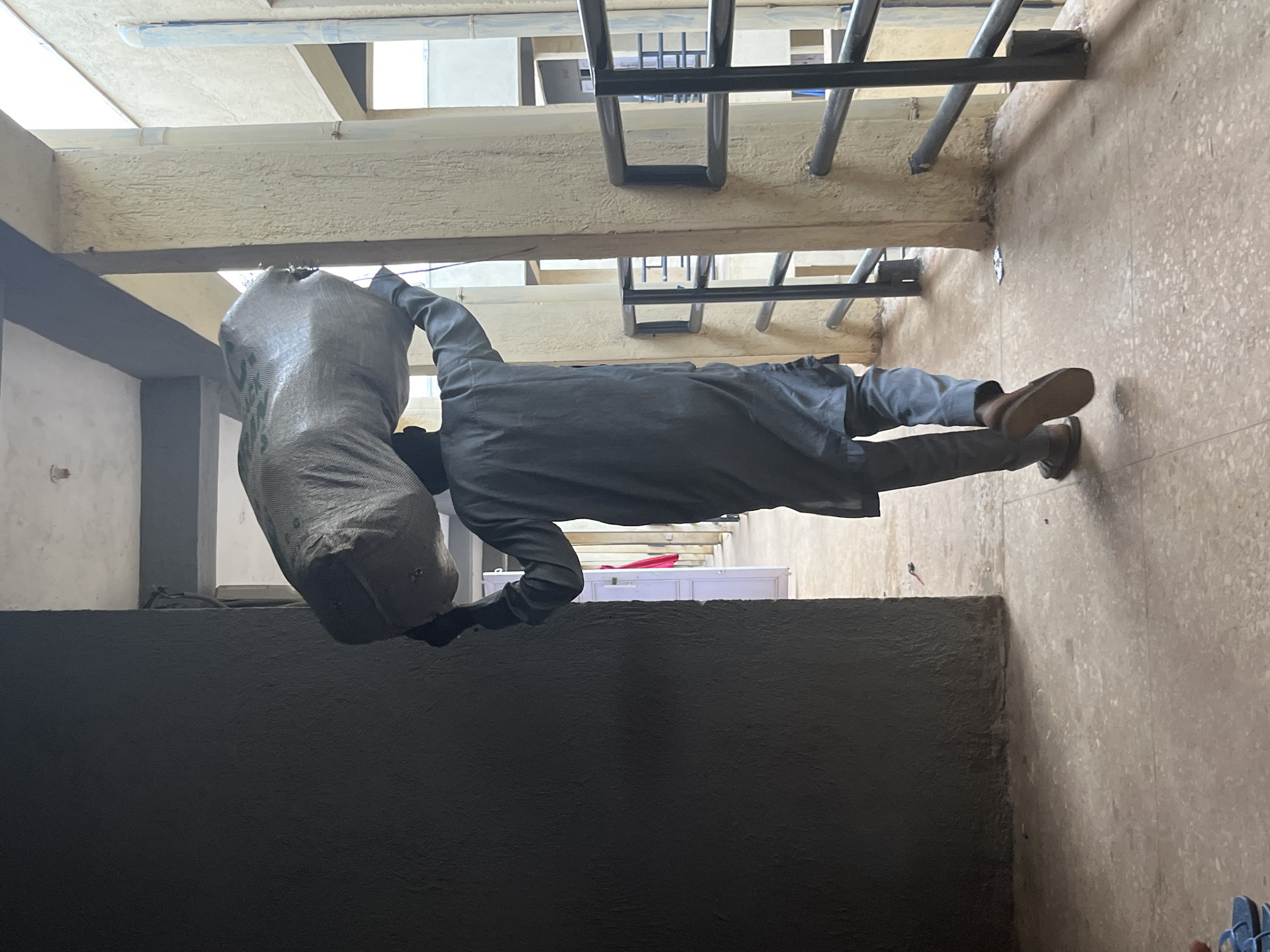 A person in a long, grey tunic and trousers walks away from the camera, carrying a large, white, woven sack balanced on their head. The person is walking down a covered walkway with a textured, beige pillar on the right and a dark, rough-textured wall on the left. The walkway has a speckled, reddish-brown floor. In the background, through the open spaces between pillars, one can see a multi-story building with balconies and railings. A bright pink cloth hangs from an opening in the wall to the left, suggesting it might be a doorway or a room. The lighting suggests it is daytime, with some shadows cast on the walkway. The scene appears to be outdoors in a built environment, possibly a market, residential complex, or commercial area in Jos, Nigeria, where such overhead carrying of goods is common.
