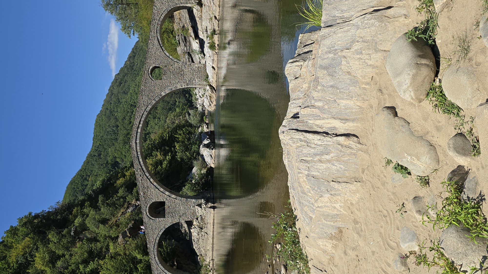 The image depicts the Devil's Bridge, a medieval stone arch bridge spanning the Arda River in the Rhodope Mountains near the village of Dyadovtsi, Bulgaria. The bridge is characterized by its three large arches and a smaller arch, all constructed from massive stone blocks. The river below is calm and reflects the bridge and surrounding lush green hillsides. The foreground is composed of sandy ground with scattered rocks and sparse vegetation, suggesting a natural, somewhat rugged environment. The sky is a clear, bright blue. Two figures are visible standing on the top of the bridge's parapet, seemingly observing the view.