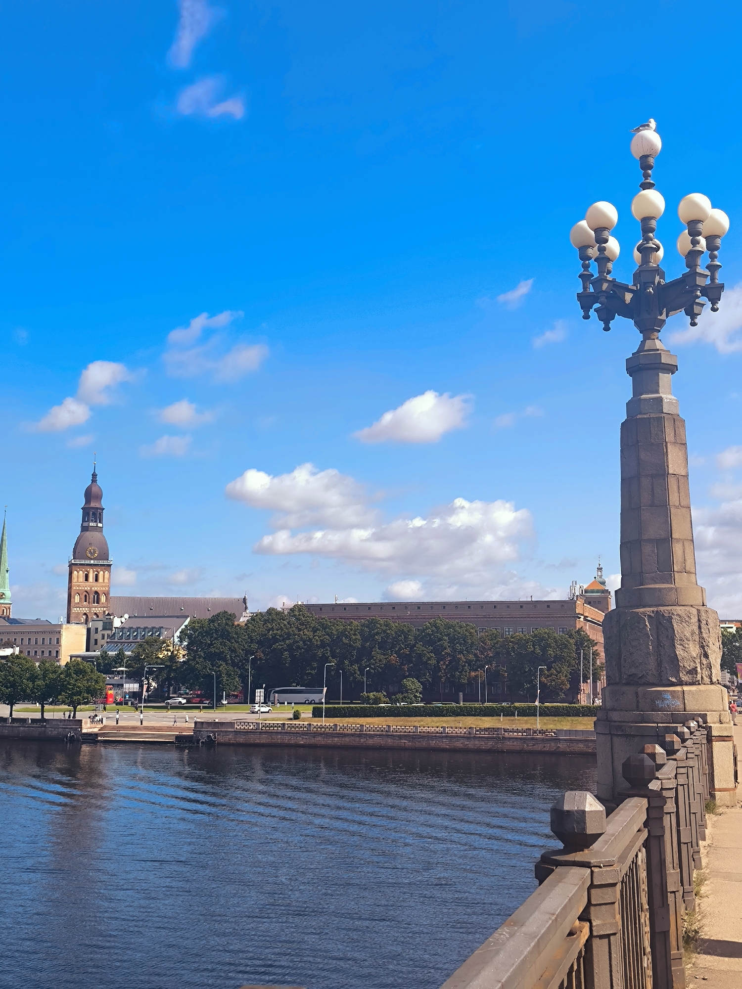 The image captures a bright, sunny day in Riga, Latvia, likely from a bridge overlooking the Daugava River. In the foreground, a stone lamppost with multiple lights stands prominently, topped with a seagull. A dark metal railing extends along the right side of the image, separating a sidewalk from the river. The river itself is dark blue with rippling patterns on its surface. Across the river, a cityscape is visible. The most notable structure is the Riga Dome Cathedral, with its distinctive tall, brown spire and clock face. Adjacent to the cathedral are other buildings with classical architecture, and a lush line of green trees separates the buildings from the riverfront. A road with pedestrian pathways, benches, and lampposts runs along the embankment. Several people are seen walking or standing in the background, and vehicles, including a white bus, are present on the road. The sky is a vibrant blue, dotted with scattered white clouds.