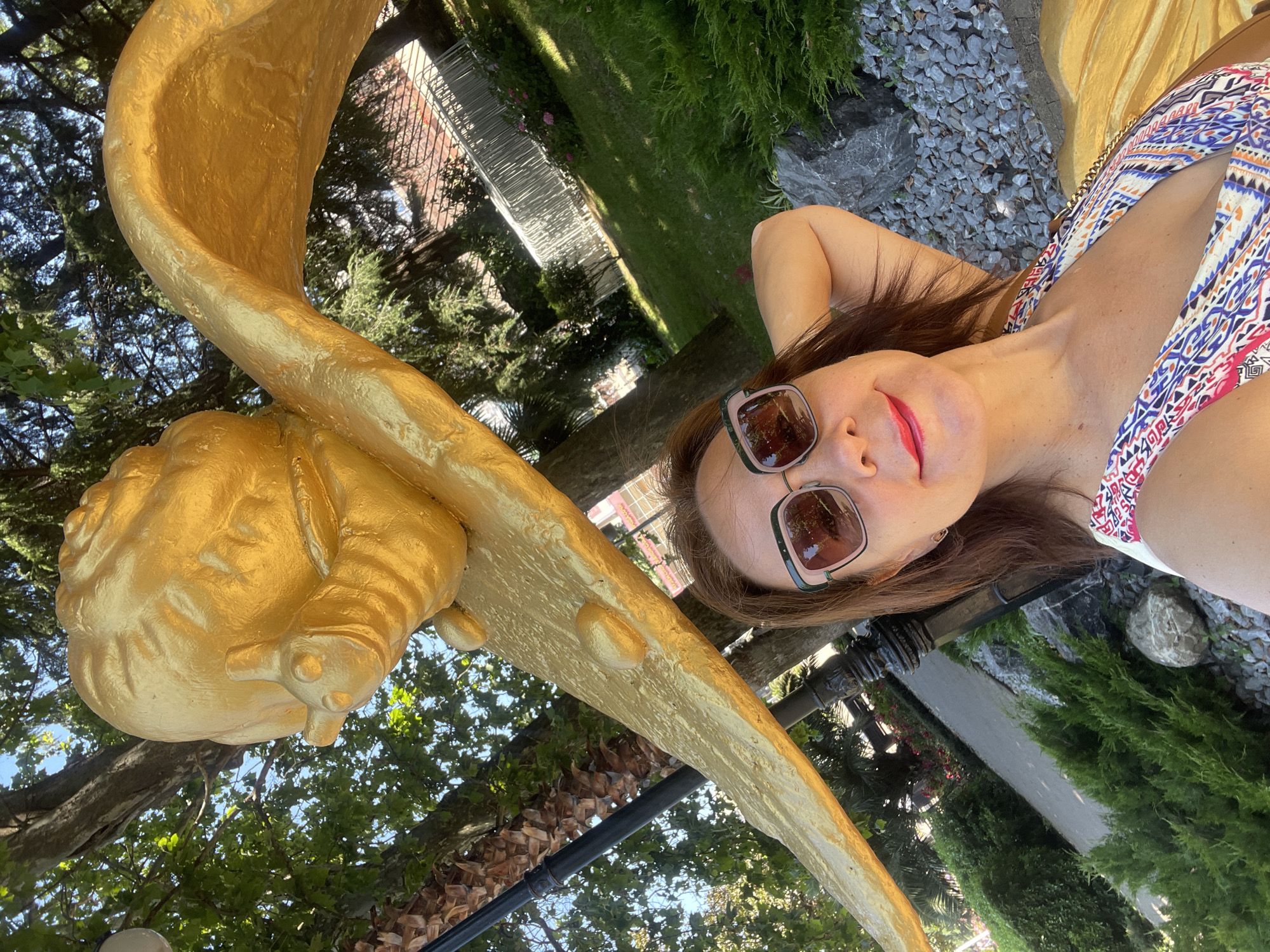 In this media file, a woman is captured in a selfie outdoors, lying on her back. She is wearing a colorful patterned bikini top and sunglasses. Her hands are behind her head, and she is smiling. To her left is a large, golden sculpture, possibly an angel or mythological figure, which takes up a significant portion of the frame. The background shows lush green trees, gravel, and hints of a building, suggesting a garden or park setting. The lighting indicates it is daytime, likely sunny. The overall mood is relaxed and cheerful. While the exact location is not specified in the image, the provided context places it in Sochi, Russia. There is no visible text in the image.