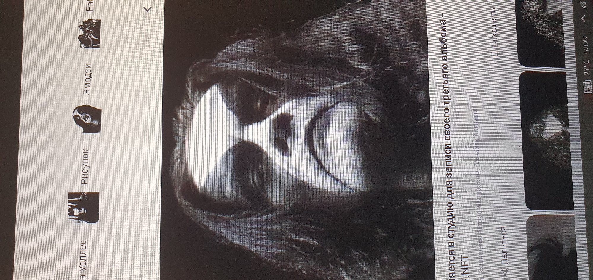 A black and white, close-up, headshot photo shows a person with black hair and white face paint. The paint is applied in a dramatic, theatrical style with broad white strokes across the forehead and cheeks, creating a stark, mask-like appearance. The person's eyes are dark and appear to be looking directly at the camera, with the white paint accentuating the orbital area. The lighting creates strong shadows, adding to the intense and possibly menacing mood of the portrait. The background is dark and out of focus, further emphasizing the subject. This image is displayed on a screen, evidenced by the horizontal lines that are characteristic of display technology. Below the main image, a line of text in Russian reads, "записи своего третьего альбома -" which translates to "recording of his third album -". Further text below this is partially visible. There are also icons and text elements suggesting a user interface for a website or application. The image is part of a screenshot that also captures a corner of the Bat Yam, Israel city screen. The temperature displayed at the bottom of the screen is 27°C.