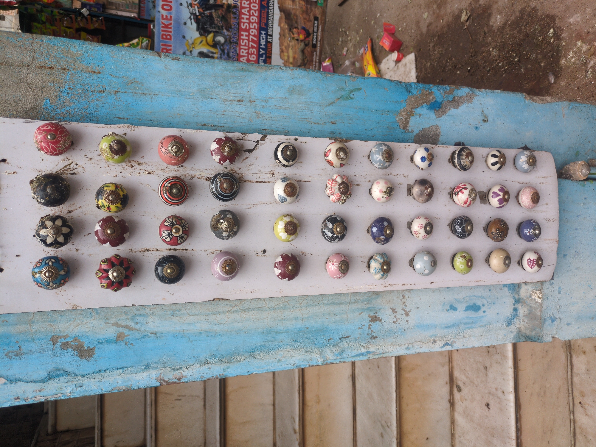 In Jodhpur, India, a display of decorative ceramic drawer knobs is mounted on a weathered, blue-painted pillar. The knobs, each approximately 2 inches in diameter, are arranged in a grid of six columns and seven rows. They feature a variety of designs, including floral patterns, geometric motifs, and solid colors such as red, blue, green, pink, and yellow, often with intricate metallic accents. Some knobs show signs of wear. To the left of the pillar, a staircase with light-colored steps is visible. To the right, a portion of a colorful advertisement for "RI BIKE ON" and "MEHRANGARH" is displayed, along with some scattered litter on the ground, including red boxes and yellow plastic packaging. The overall scene suggests a street-side stall in a bustling urban environment.
