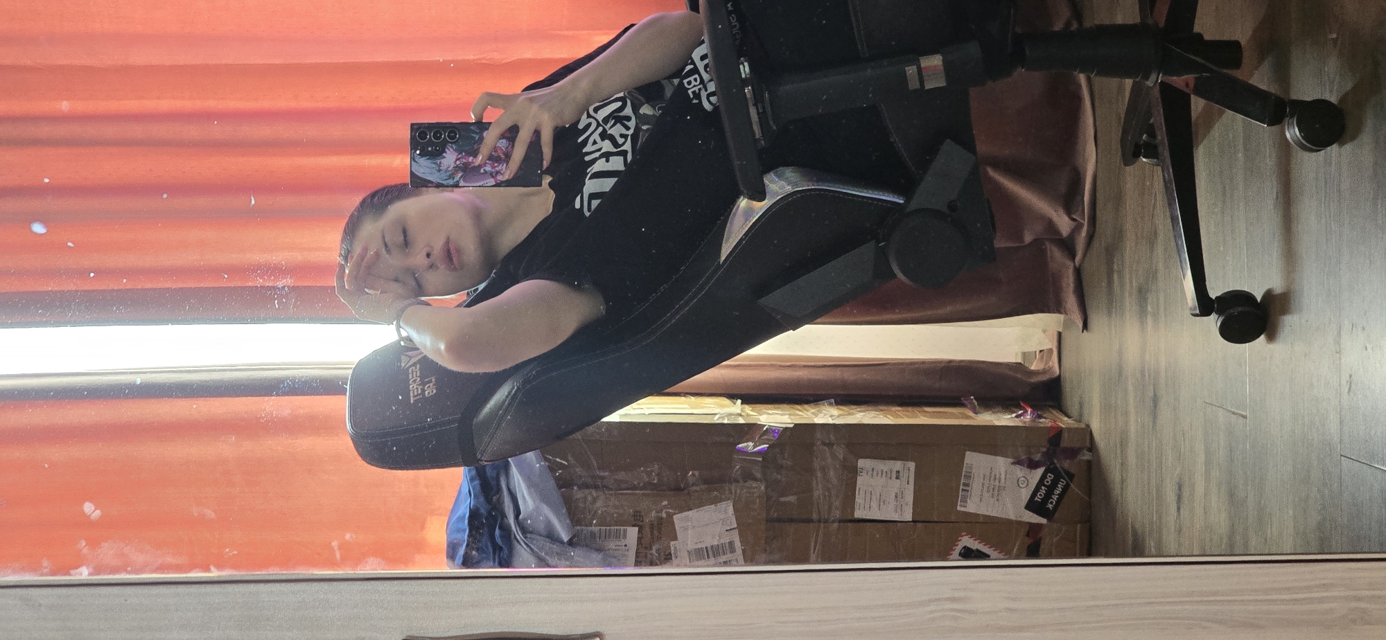 The image is a mirror selfie taken by a young woman indoors, reflected in a full-length mirror. She is sitting in a black gaming chair, leaning back with her right arm raised and hand resting on her forehead. She is wearing a black t-shirt with a graphic print. In her right hand, she holds a smartphone, which is angled to capture the selfie. The phone case has an anime-style illustration.
The background visible in the mirror includes a floor with a wood-like pattern, several cardboard boxes stacked and wrapped in plastic, and orange curtains partially drawn. The boxes appear to be moving or storage boxes, with shipping labels and tape visible. A portion of a wooden closet or cabinet is visible on the left edge of the frame. The lighting suggests an indoor environment, possibly a room in an apartment or house. There is no information in the image that indicates a specific location in the city of Burgas, Bulgaria.