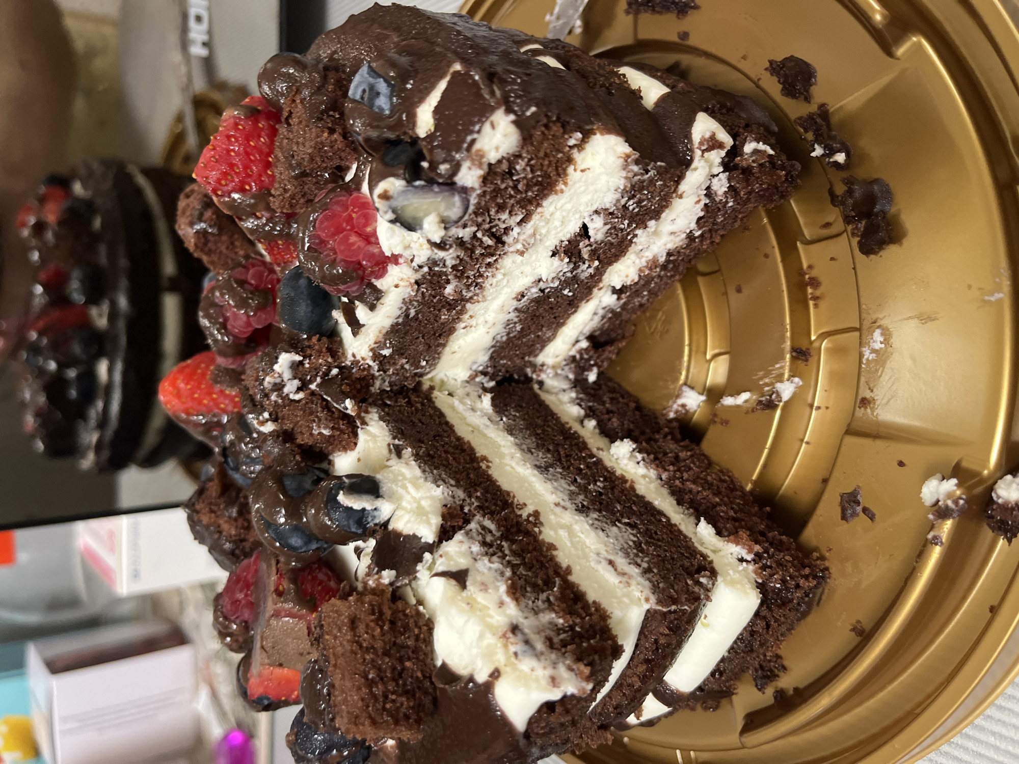 A slice has been taken from a multi-layered chocolate cake topped with whipped cream, chocolate frosting, chocolate chunks, strawberries, blueberries, and raspberries. The cake is situated on a gold-colored plastic platter. In the background, a blurred second chocolate cake is visible, also adorned with berries. The scene appears to be indoors, possibly in a kitchen or dining area, given the presence of what looks like a laptop and boxes in the background. No individuals are clearly depicted in the image. The environment is indistinct beyond the immediate tabletop area. There is no information within the image to suggest it is located in Sochi, Russia.