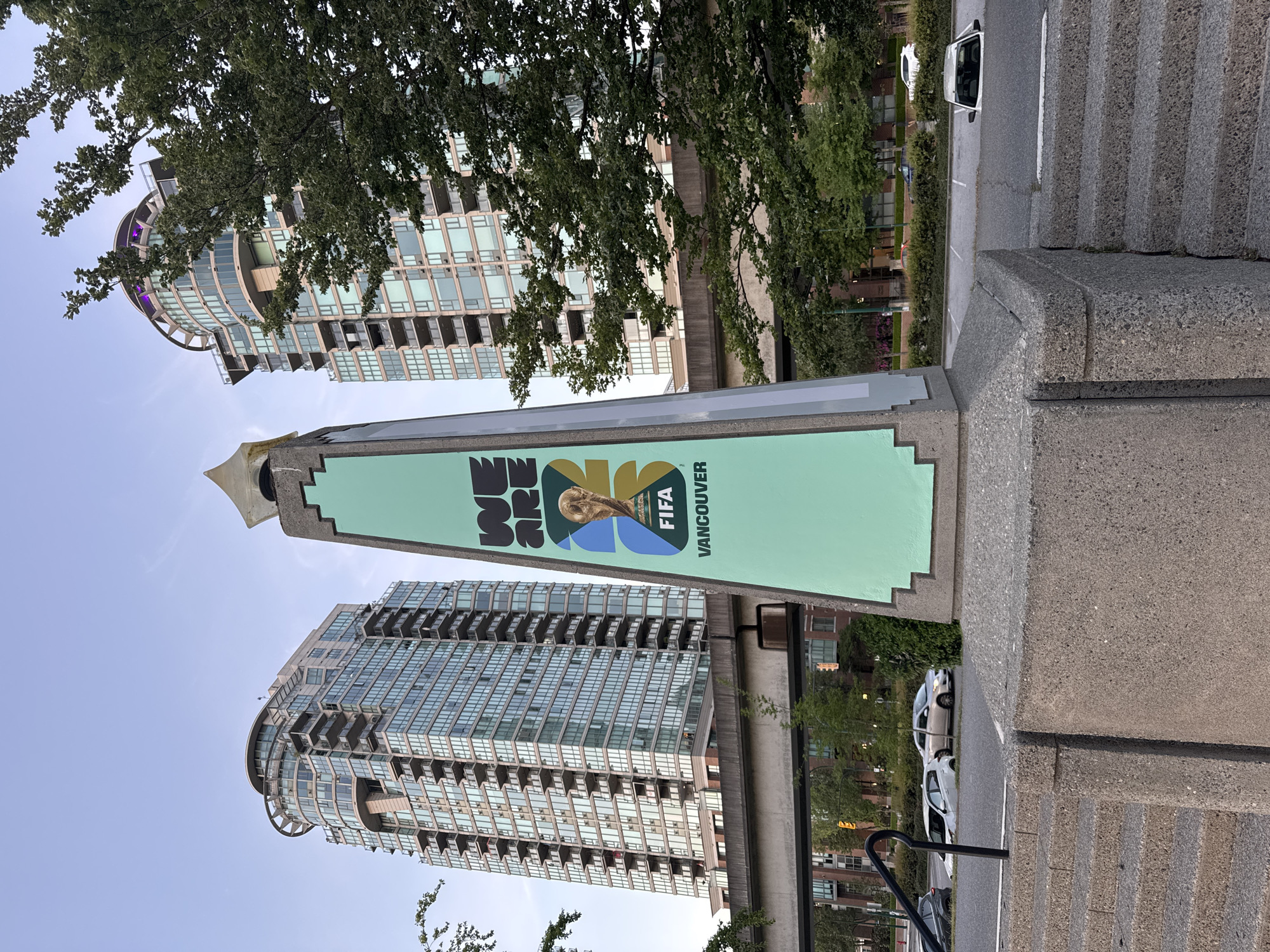 The image captures a street scene in Vancouver, Canada. Dominating the foreground is a tall, green sign promoting the FIFA 2026 World Cup, with "Vancouver" clearly visible on the sign. Behind the sign are two modern, high-rise residential buildings featuring glass facades and rounded architectural details. A few trees are visible, partially obscuring parts of the buildings. To the right of the sign are some concrete stairs leading down to the street, with parked cars visible. The sky is light blue and mostly clear. The image is taken from a slightly low angle, emphasizing the height of the sign and buildings.