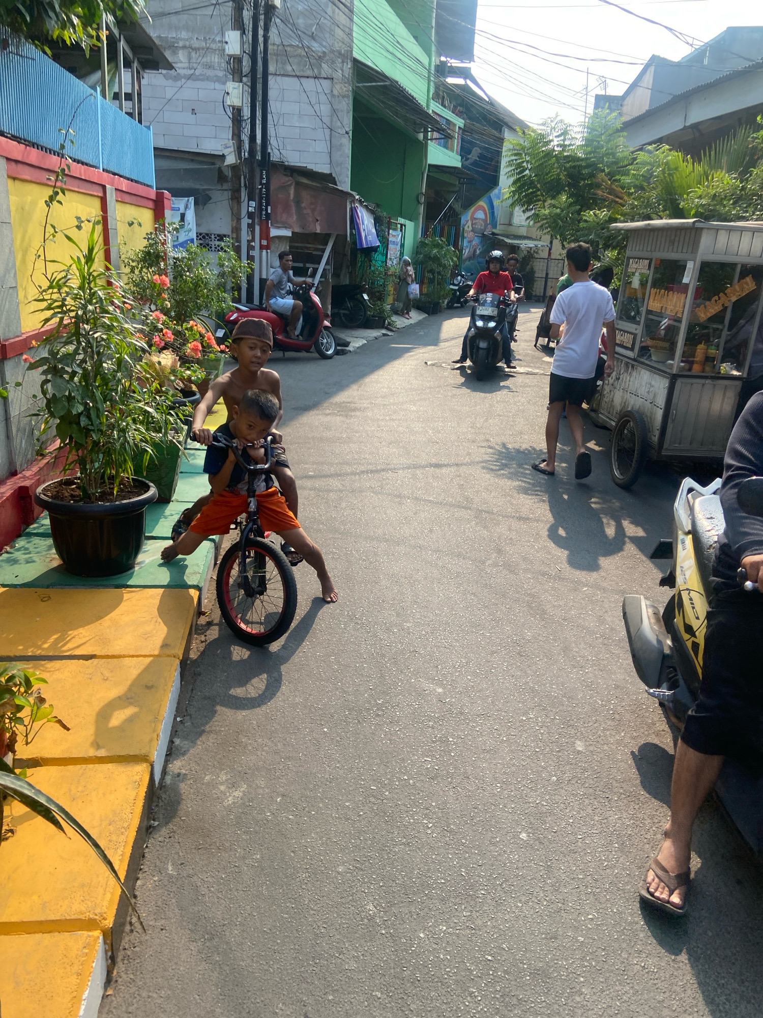 In a narrow, sunlit street in North Jakarta, Indonesia, two young boys are the central focus. The younger boy, wearing orange shorts and a dark t-shirt, sits on a small black bicycle with red rims, his feet dangling. He is positioned on the road, slightly to the right of center. His older brother, wearing a brown cap and no shirt, sits behind him on the bicycle, his arms around the younger boy. They appear to be in motion or preparing to move.
To the left of the boys, a large dark pot with a leafy green plant sits on a yellow-painted concrete curb. The curb extends to the left edge of the frame. Behind the curb is a building with a yellow lower facade and a blue metal fence above it.
Further down the street, on the right side, a food cart with a glass display case and various bottles is parked. To the left of the food cart, several people are visible. A man in a white t-shirt and black shorts is walking away from the camera. A person on a motorcycle, wearing a red shirt and helmet, is also in motion, heading away from the viewer. Another motorcycle is visible further back.
The road surface is asphalt, with visible cracks and texture. The scene is illuminated by bright sunlight, casting distinct shadows on the road. Buildings line both sides of the street, some with corrugated metal roofs and others with a more modern facade. Power lines crisscross overhead. The overall impression is of a busy, everyday scene in a densely populated urban environment.