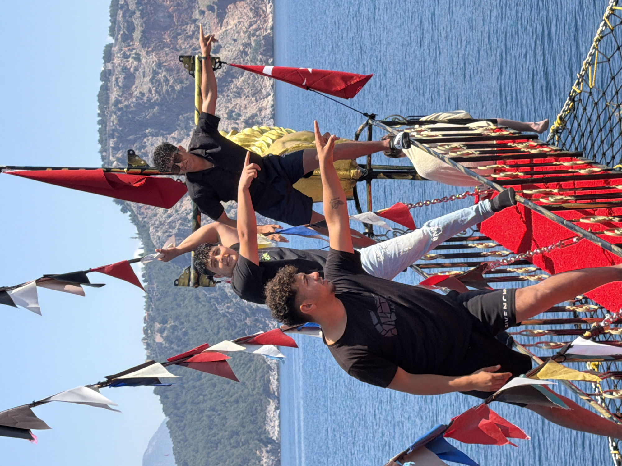 The image captures four young men posing playfully on a boat, likely a pirate ship replica, against the backdrop of a clear blue sky, calm blue sea, and a mountainous coastline, consistent with Alanya, Turkey. The photo is taken during bright daylight, suggesting sunny weather.
The subjects are positioned at different levels on the boat's rigging and decks. In the lower-right foreground, a man with curly dark hair, wearing a black t-shirt and black shorts with "GIVENCHY" visible on them, extends his right arm upwards with his index finger pointing. He looks to his left. Behind him and slightly to the left, another man, also with dark curly hair and a black t-shirt, smiles towards the camera, mirroring the upward-pointing gesture with his right hand.
Further up and towards the center, a third man in a black t-shirt and dark shorts, wearing sunglasses, raises his left arm with his index finger pointing skyward. On the far right, a fourth man, wearing a light-colored t-shirt and light blue jeans, is partially visible, appearing to relax on a woven net structure of the boat.
The boat is adorned with numerous colorful pennant banners in red, white, black, and blue. Several larger red flags are also present, one of which prominently features a white star and crescent, indicating it is a Turkish flag. The boat's structure features red-painted elements, metal railings, and black and yellow netting. The background shows rugged, rocky mountains covered with sparse greenery, meeting the tranquil blue sea under a cloudless sky. The overall mood is one of enjoyment and recreation.