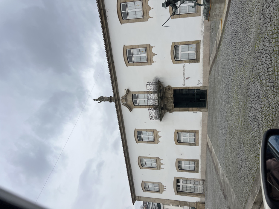 An exterior shot depicts the Câmara Municipal (Town Hall) of Santa Marta de Penaguião, Portugal, under an overcast sky. The prominent two-story building features a white painted facade with numerous rectangular windows trimmed in light-colored stone, each fitted with white frames and internal light-colored curtains. A central, ornate stone balcony with a black wrought-iron railing sits above the main entrance, which is dark and appears open. A stone statue of a robed figure, possibly religious, stands atop the roof directly above the balcony. The building's brown tile roof is visible. "CÂMARA MUNICIPAL" is inscribed in dark lettering on the white wall to the right of the main entrance. The foreground is a large cobblestone square. To the far right, a partial view of a bronze-colored statue on a stone pedestal shows a figure wearing a hat and holding a long-handled tool over his shoulder. In the lower-left corner, a car's side mirror is visible, reflecting a portion of the vehicle's interior and a person's face. Hills are discernible in the distant background on the left. A thin dark wire traverses the upper portion of the frame horizontally.
