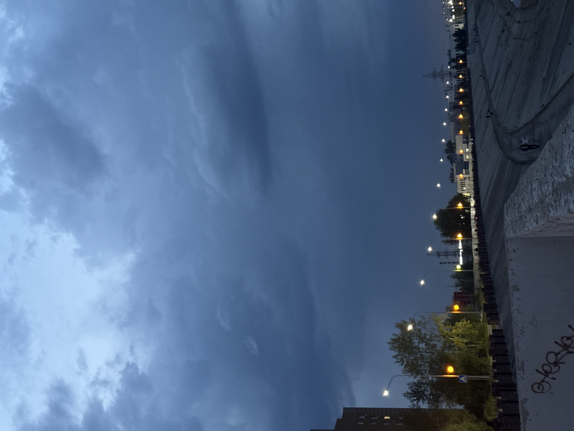 The image depicts a twilight scene, likely on the embankment of the Ufa River in Ufa, Russia. A dense, dark cloud cover dominates the sky. In the foreground, a concrete barrier with graffiti is visible. Beyond this, a sloped, paved area lines a body of water, possibly the river. A path winds along this embankment, illuminated by streetlights. Several figures can be seen walking along the path. On the opposite side of the embankment, trees and buildings, including a tall brick structure on the left, are present. Industrial elements, such as power lines and towers, are also visible in the distance. The lighting suggests the time is either dusk or dawn, with artificial lights from streetlamps and buildings adding to the illumination.