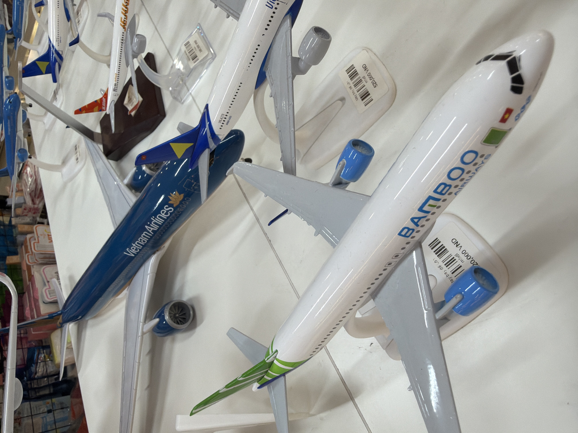 The image depicts a collection of model airplanes displayed on a white surface, likely a tabletop in a retail setting in Đà Nẵng, Vietnam. The models represent various airlines, including a prominent blue Vietnam Airlines plane with its branding clearly visible. To the left, a white airplane with green accents and "BAMBOO AIRWAYS" text is in the foreground. Other models are visible in the background, some with orange and blue livery. The models are mounted on white stands. In the background, out-of-focus shelves stocked with merchandise are discernible, suggesting a shop or market environment. The lighting is bright and even, typical of indoor retail spaces. There are no people or animals visible in the frame.