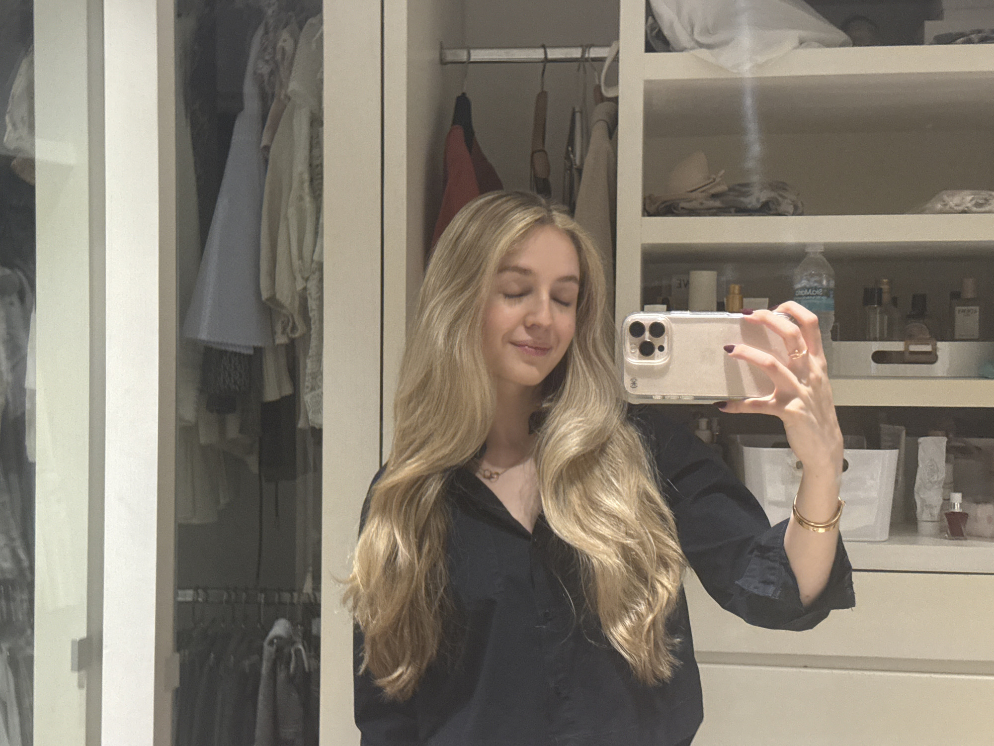 A young woman with long, wavy blonde hair, wearing a black shirt and gold bracelet, is taking a selfie in what appears to be a walk-in closet. The closet has clothing hanging on racks and shelves filled with various items, including toiletries and accessories. The precise location within Mexico City, Mexico, is indeterminable without additional information.