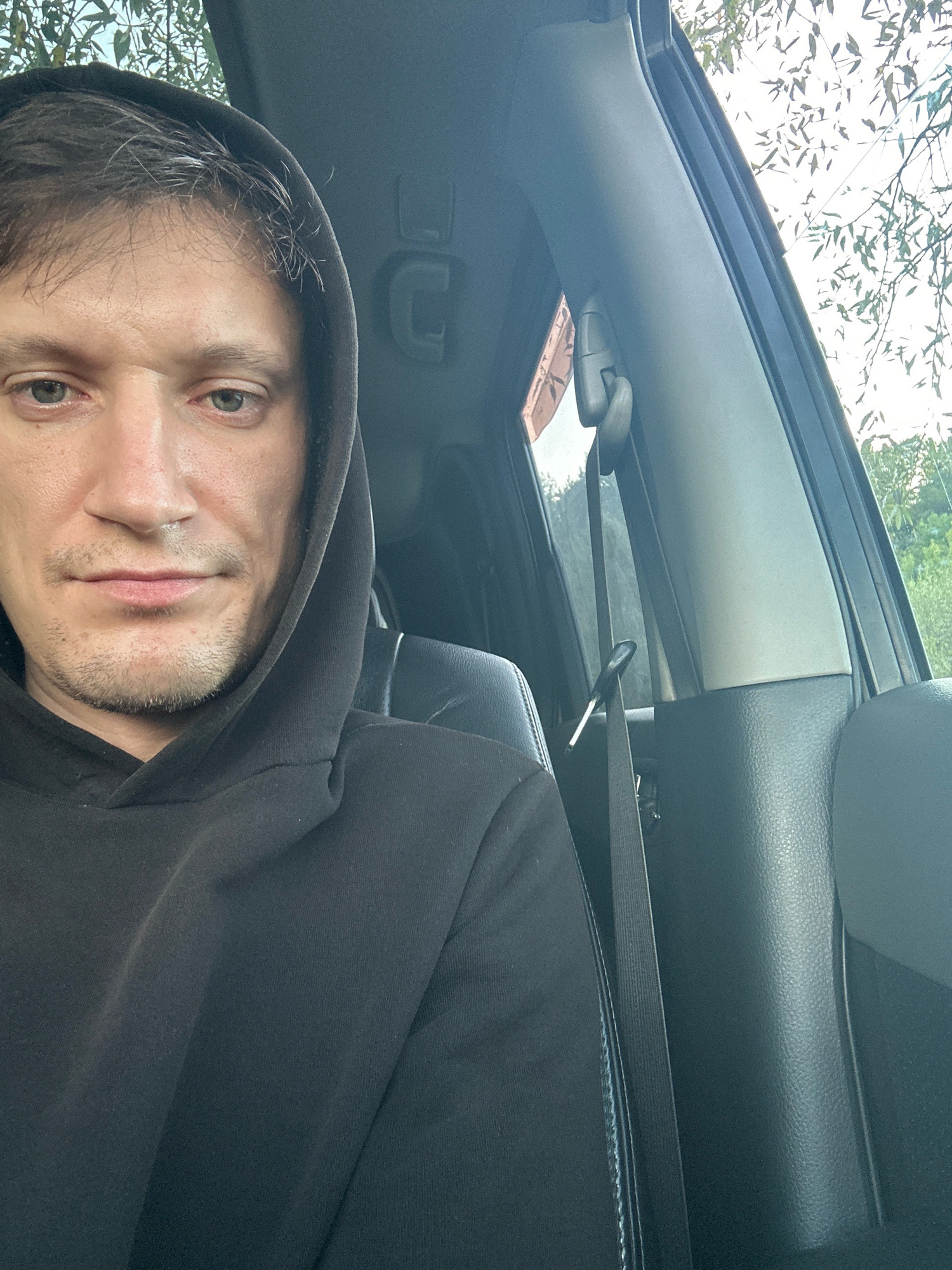 A man wearing a black hoodie is seen from the chest up, inside a car. He is looking towards the camera, with his head slightly tilted. The car's interior is visible, including a black seat and part of the car door. Outside the car, through the window, foliage and a bright sky can be seen. The scene appears to be captured during daylight. There is no specific indication of the location being Makiv, Ukraine.