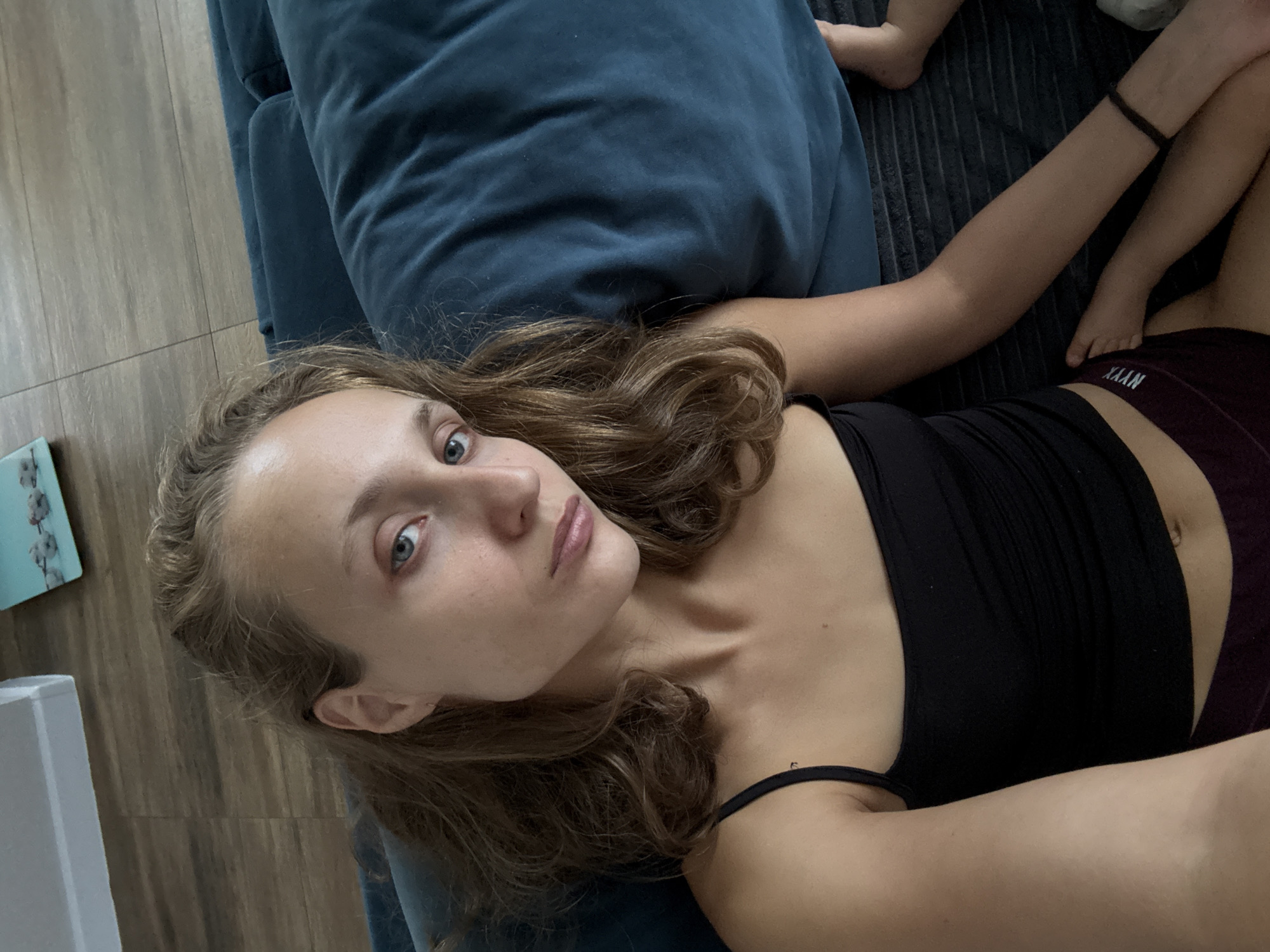 This is a selfie of a young woman lying down on a dark couch with a blue pillow. She is wearing a black tank top and burgundy shorts with the word "XYXAN" visible on the waistband. The woman has long, wavy brown hair and blue eyes. She is looking directly at the camera with a neutral expression. The scene appears to be indoors, possibly in a living room, with wooden flooring visible. The lighting suggests it might be daytime. A small portion of another person's leg, presumably a child's, is visible in the upper right corner of the image.