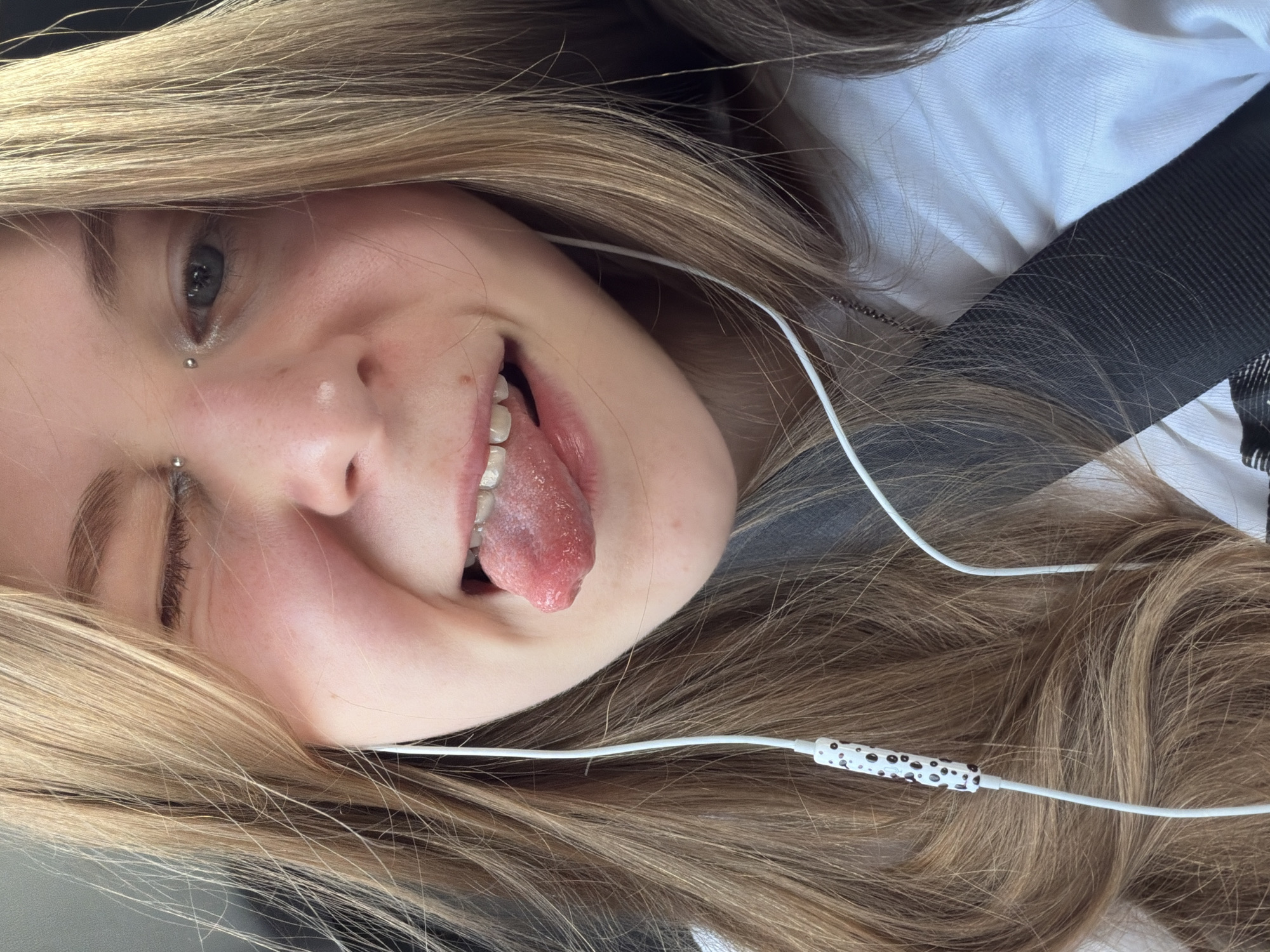 A close-up shot features a young person with blonde hair, partially obscuring their face. They are winking with their left eye and sticking out their tongue. A vertical piercing is visible on their eyebrow. They are wearing white earbuds with a patterned control piece. The person is wearing a white shirt and a black seatbelt. The background is blurred, suggesting they are in a vehicle. The image is set in the city of Novoivanovskoe, Russia.