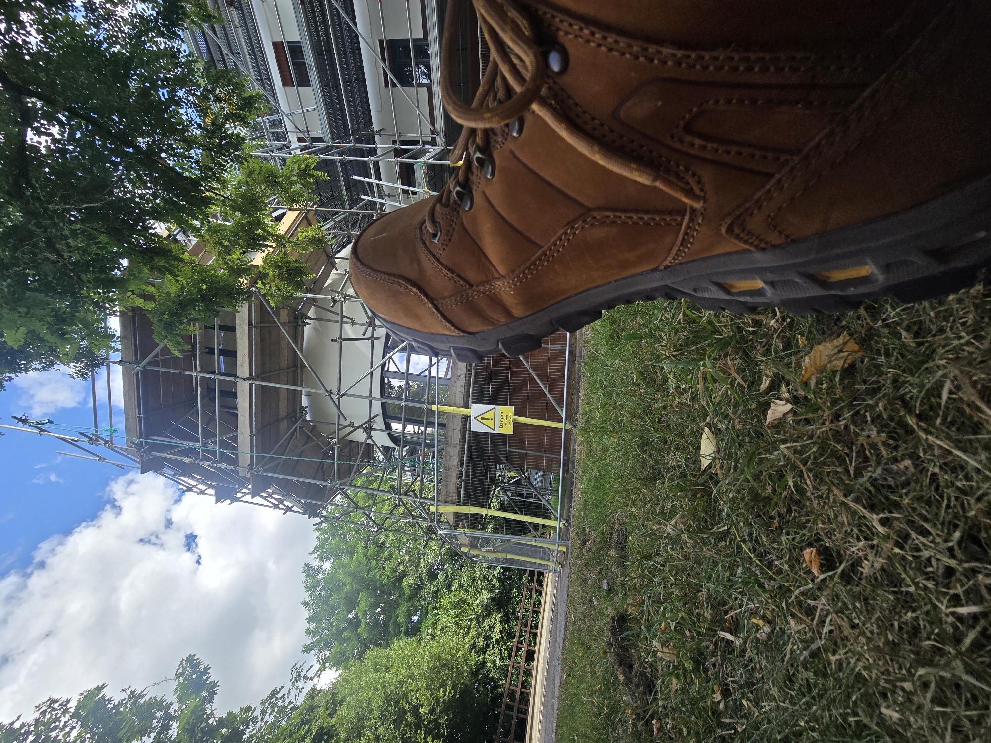 The image shows a close-up, low-angle view of a brown work boot in the foreground, resting on grass. Behind the boot, a building is under construction, covered in scaffolding. A yellow warning sign is visible on the scaffolding, indicating "Danger: Workers and Public." The environment appears to be outdoors, with trees and a blue sky with clouds visible. The scene is likely located in Woking, United Kingdom, given the context of construction and typical outdoor settings in that region.