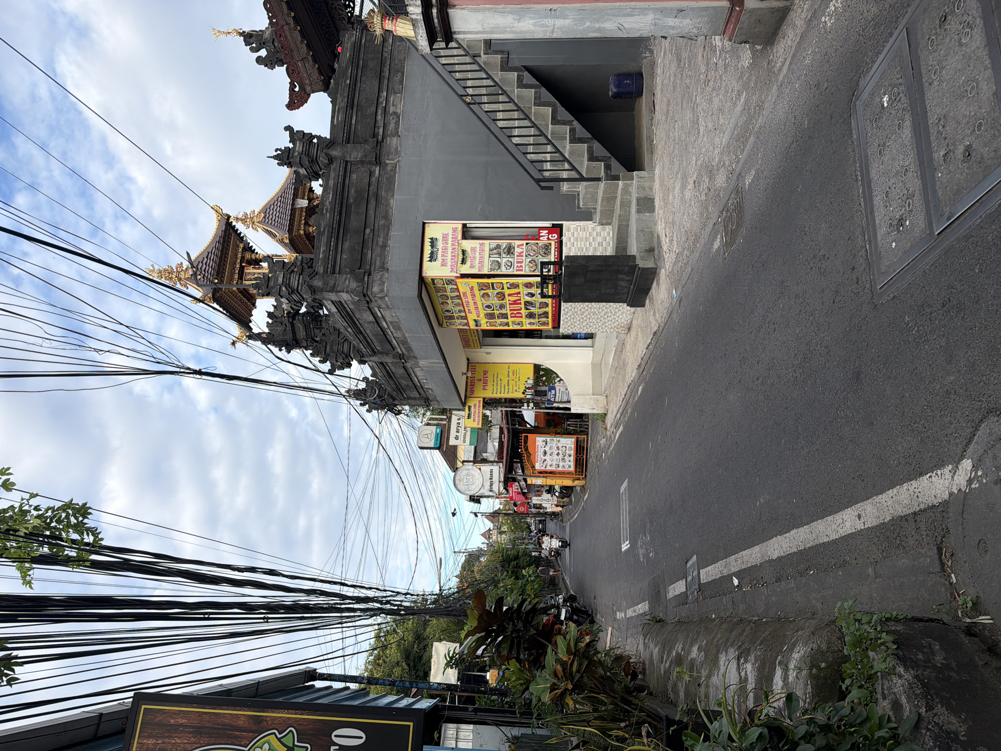 A street scene in Kuta, Indonesia, shows a narrow asphalt road curving slightly to the right. A traditional Balinese structure with a tiled roof and intricate carvings stands on the left side of the road, marked by a restaurant sign. A dense web of tangled electrical wires stretches above the street and alongside buildings, obscuring part of the sky.
On the right, the sidewalk runs alongside a building with a staircase leading to an upper level. The building features signs for local businesses, including a restaurant with menu items displayed. The road surface shows wear and tear with visible patches and faint white lane markings near the curb. Tropical vegetation is seen on both sides of the road.
In the distance, the road leads to a view of more buildings and light vehicular traffic. A few people are visible along the street.