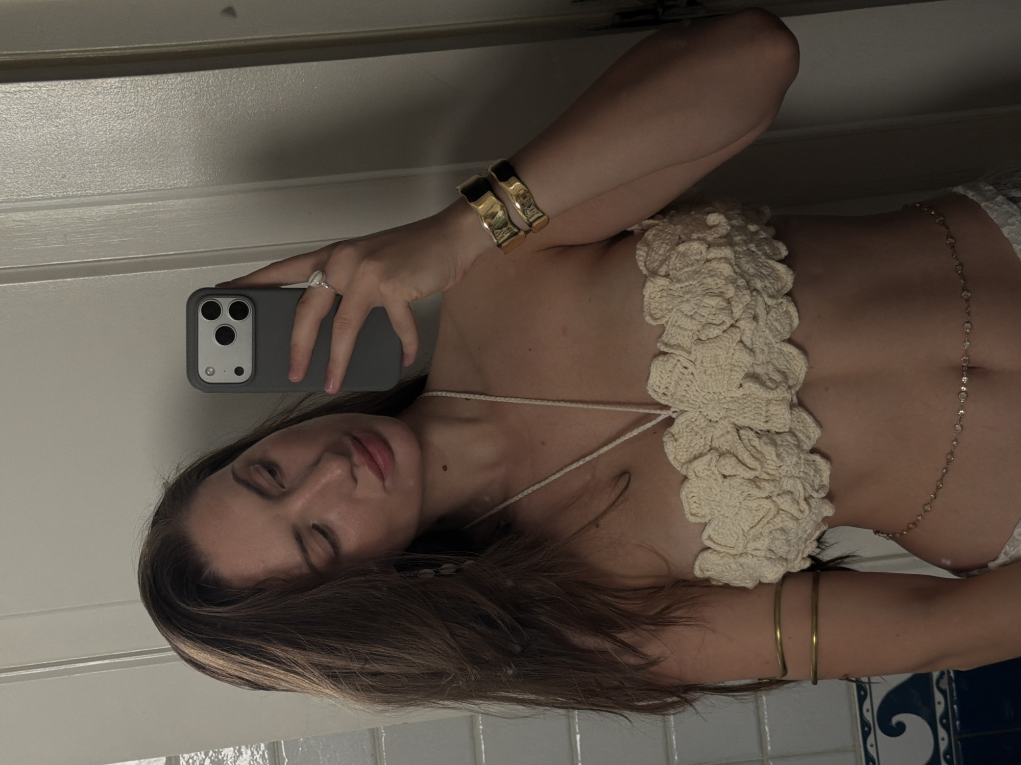 A woman is taking a mirror selfie in what appears to be a bathroom. She is wearing a cream-colored, crocheted bikini top with a floral pattern, a delicate gold chain around her waist, and two gold bracelets on her right arm. She has long, dark brown hair, and is wearing a ring on her left hand. The background shows white tiled walls and a white door. There is no indication of the city of Sharm El Sheikh, Egypt.