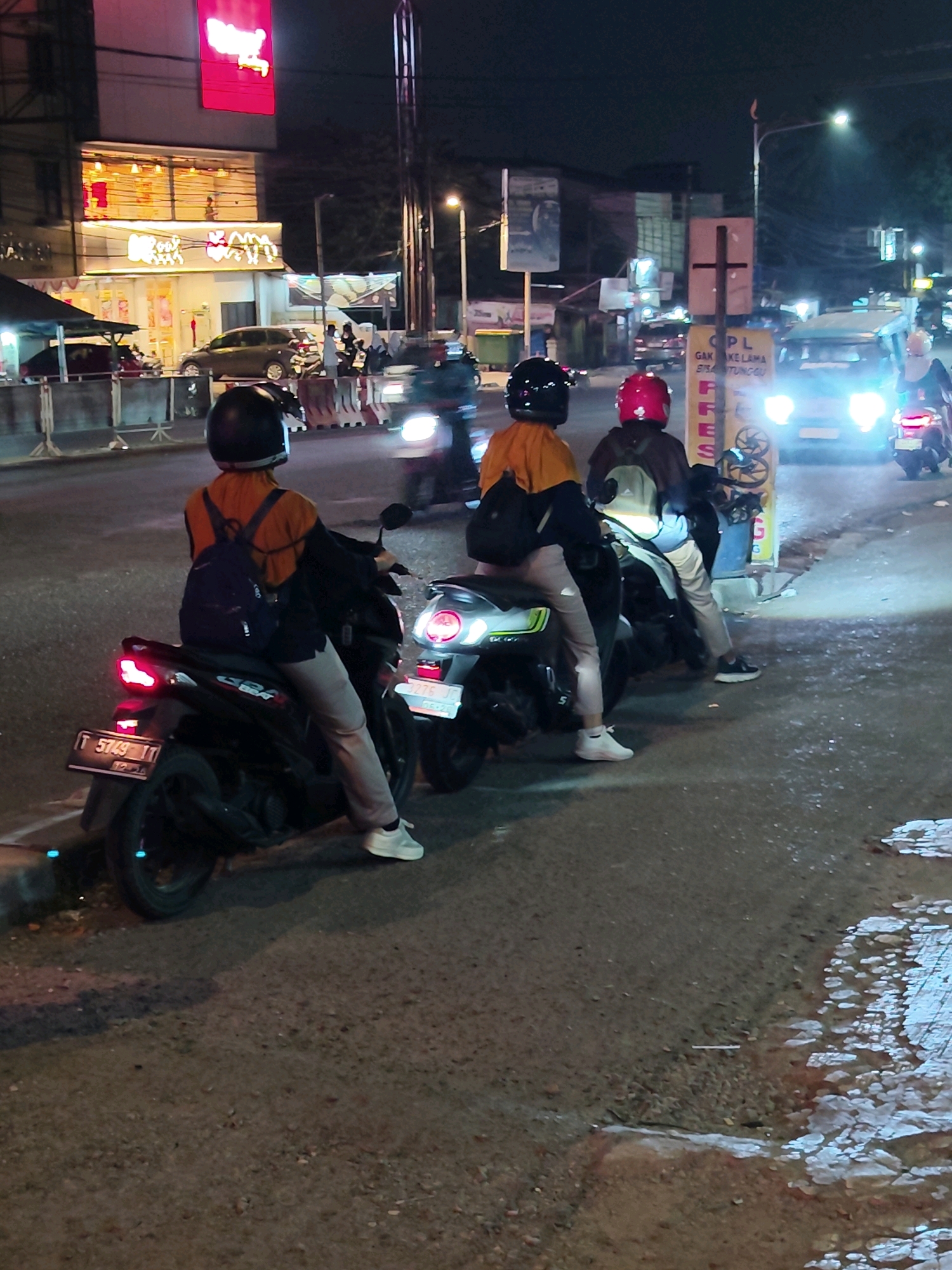 The image shows a nighttime street scene in Purwakarta, Indonesia, with a focus on several individuals on motorcycles. The setting appears to be a commercial area, with illuminated buildings and signage in the background, including what looks like a restaurant. Three people are prominently featured, seated on their motorcycles. The person closest to the foreground is wearing an orange shirt and a black helmet, with a backpack. Another person is wearing a red helmet and a dark jacket. The third individual, partially obscured, is also on a motorcycle. They are all facing the same direction, suggesting they are waiting or stopped. Several other motorcycles and a car are visible further down the street, some with headlights on, indicating ongoing traffic. The road surface is asphalt, with some debris and wet patches. The overall impression is a typical urban street environment at night in Indonesia.
