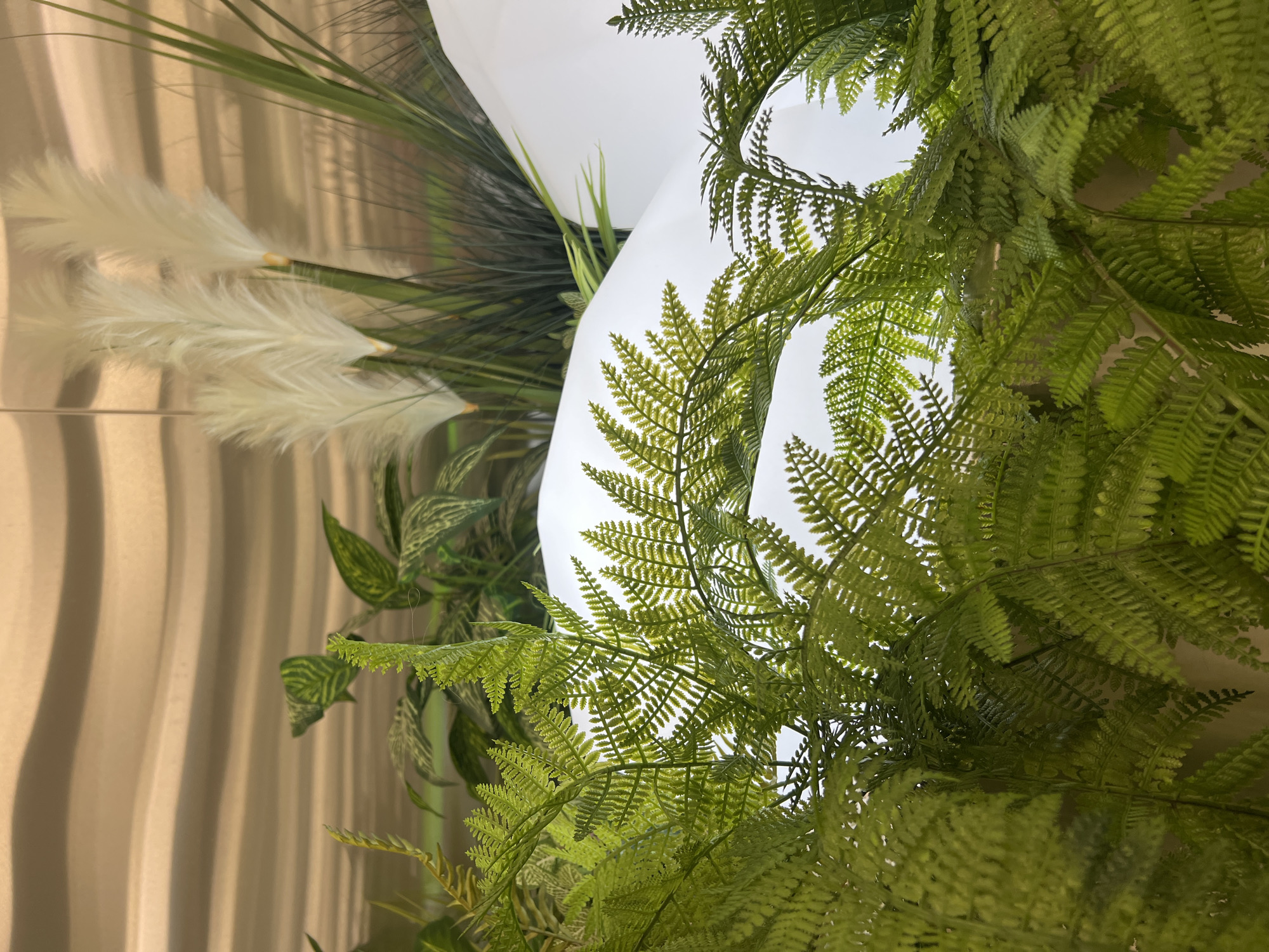 The image shows a close-up view of artificial plants, primarily ferns, in the foreground. Behind the ferns, there are white, curved surfaces that suggest a sculpted or mountainous terrain, possibly part of an interior design or a decorative display. In the background, feathery white plumes, resembling pampas grass, are visible along with some dark green, blade-like foliage. The backdrop appears to be a wall with a textured, wavy pattern in a golden-brown hue. The scene is illuminated, creating highlights and shadows on the plant materials and the white surfaces. No people or specific identifiable landmarks of Ladozhskoe Ozero, Russia are present in the image.